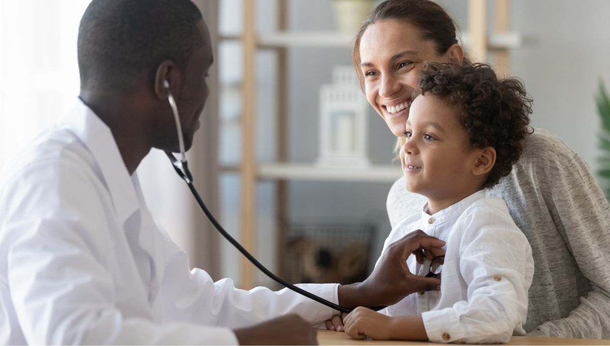 Médico examinando uma criança com um estetoscópio enquanto um dos pais sorri ao lado, em uma clínica.