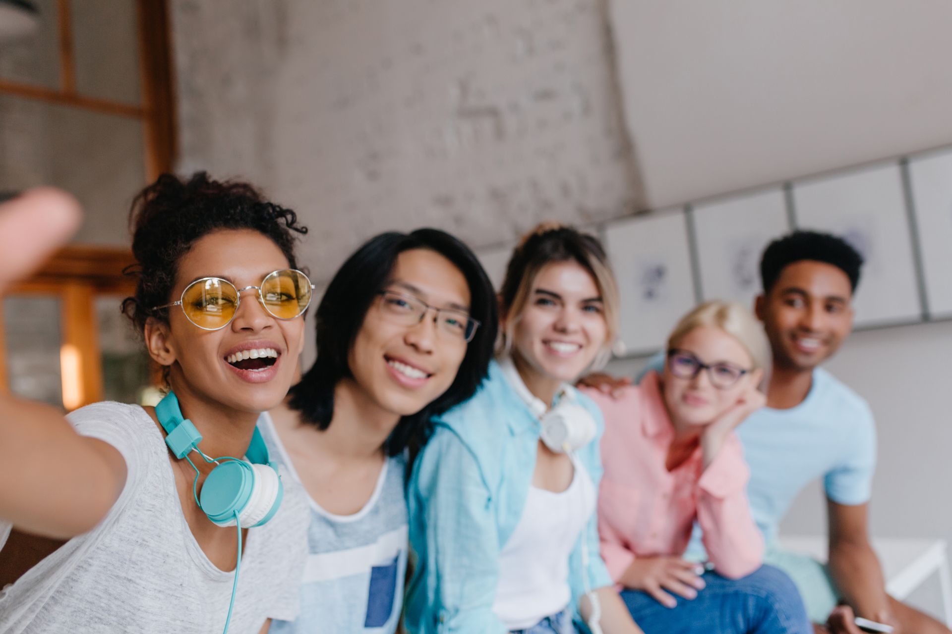 Cinco amigas sorrindo e encostadas uma na outra dentro de casa, vestindo roupas casuais coloridas.