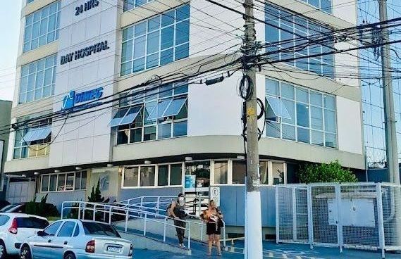 Prédio de escritórios moderno com janelas azuis, carros estacionados e algumas pessoas na entrada.