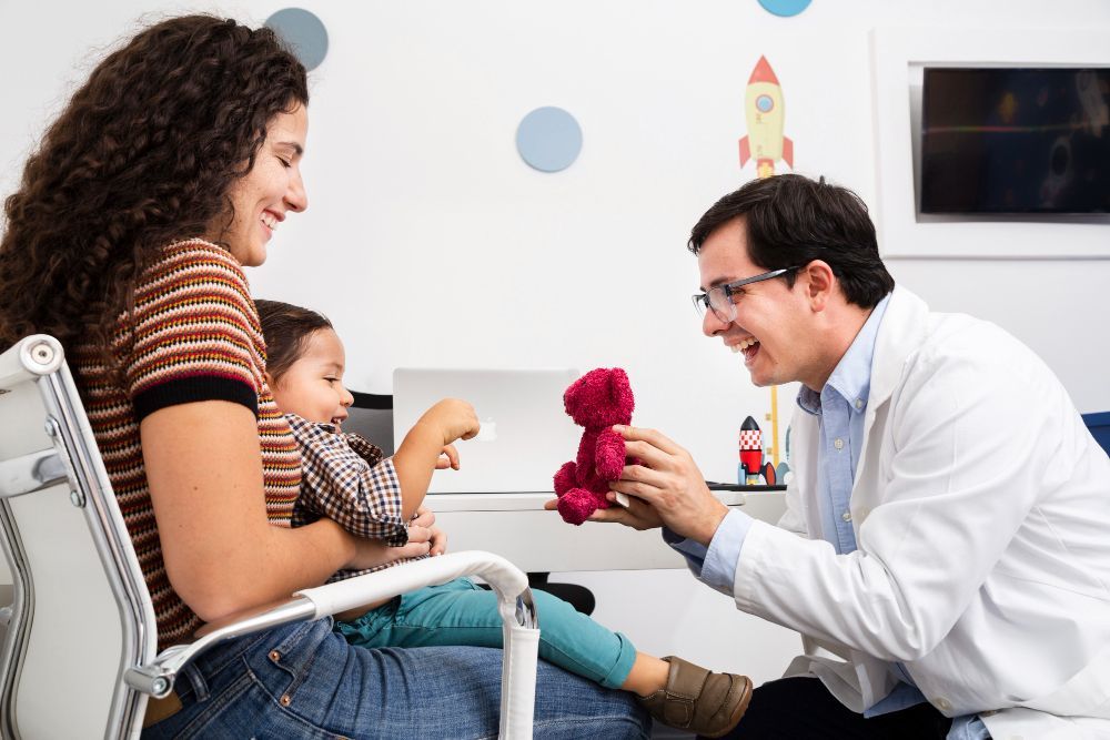 Médico ajoelhado com flores para uma mãe e seu filho sorridentes na sala de espera de uma clínica.