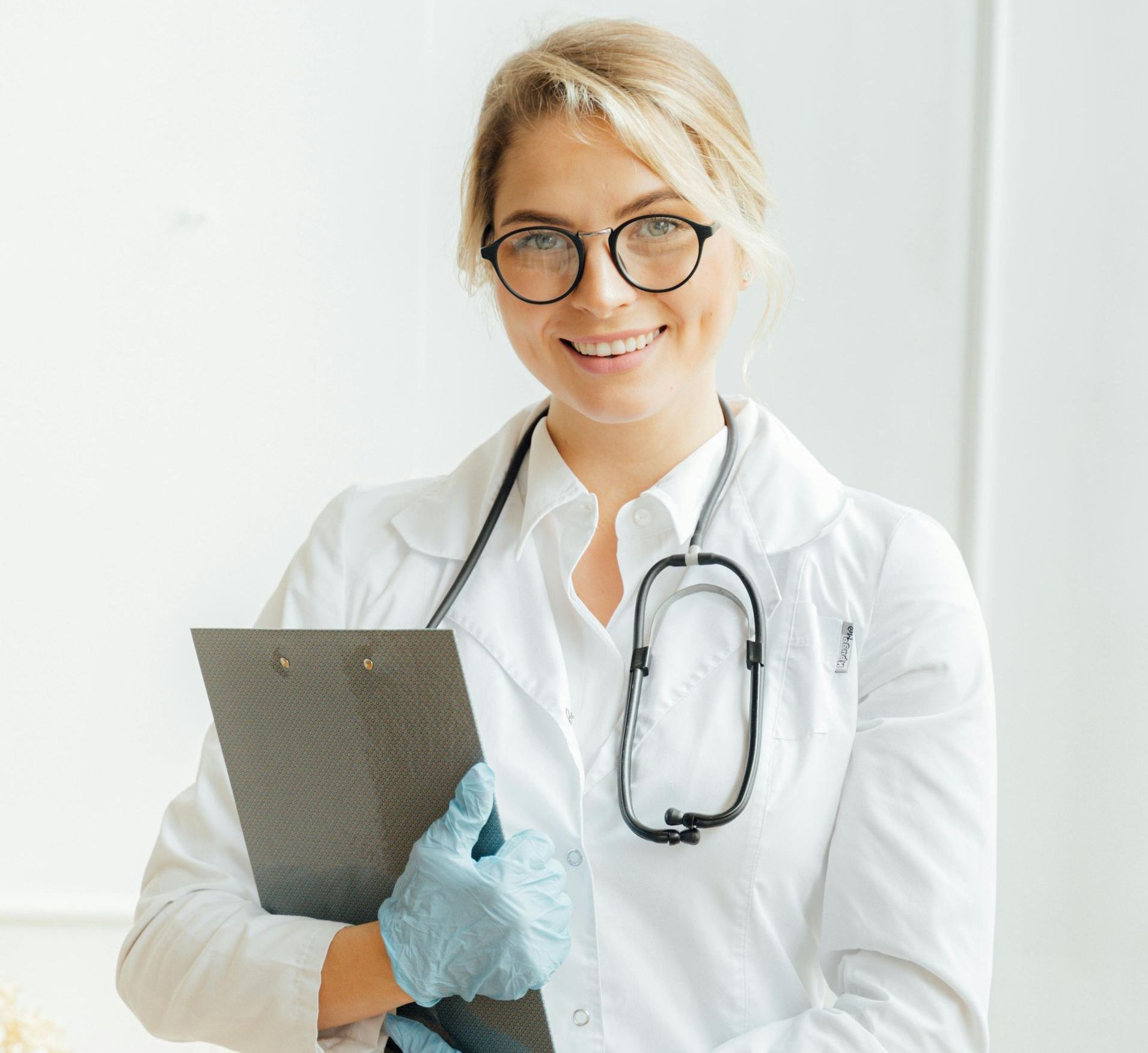 Médico sorridente segurando uma prancheta, vestindo um jaleco branco e com um estetoscópio em uma clínica bem iluminada.