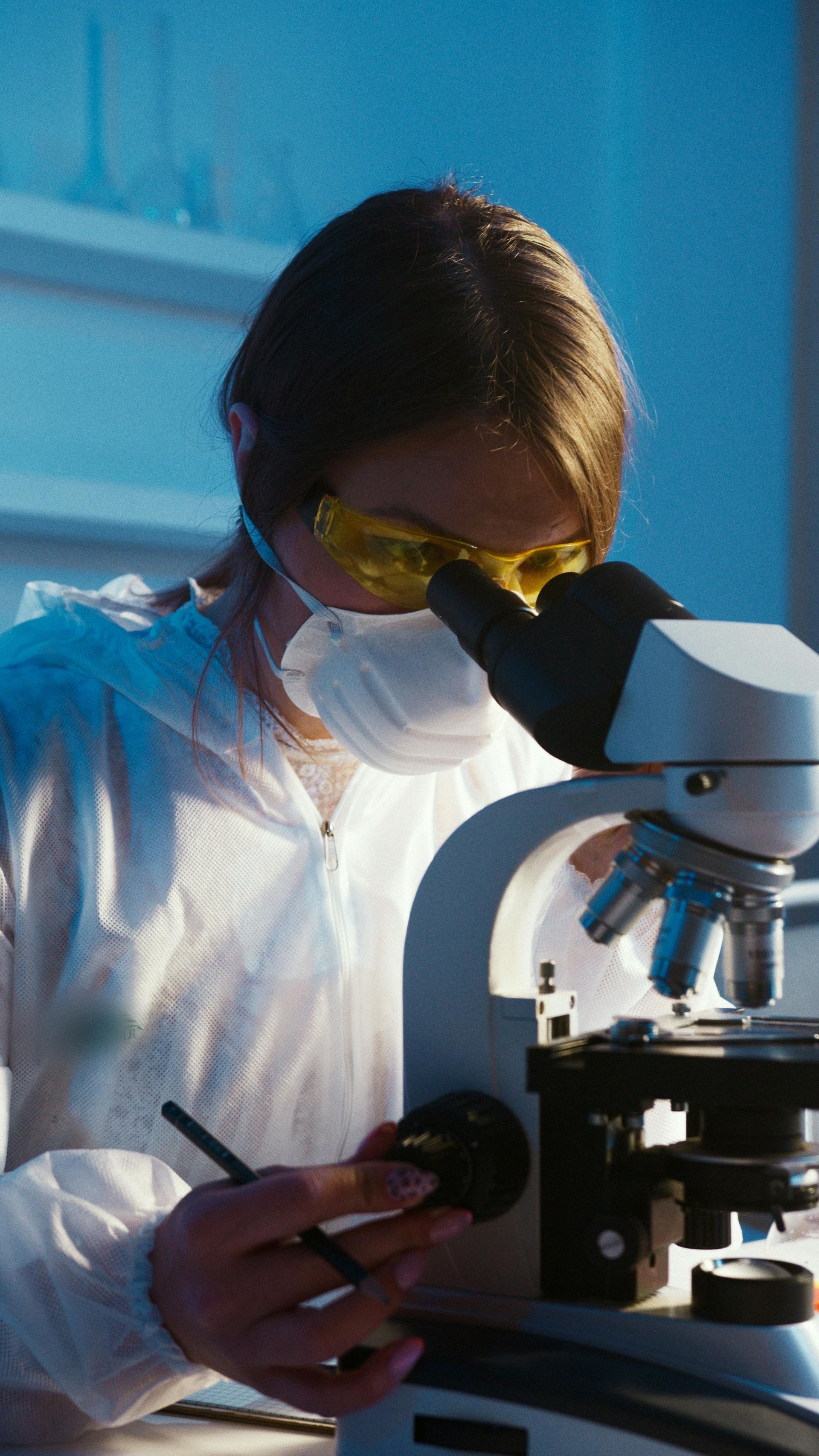 Cientista de jaleco branco e máscara examinando uma amostra ao microscópio em um laboratório.