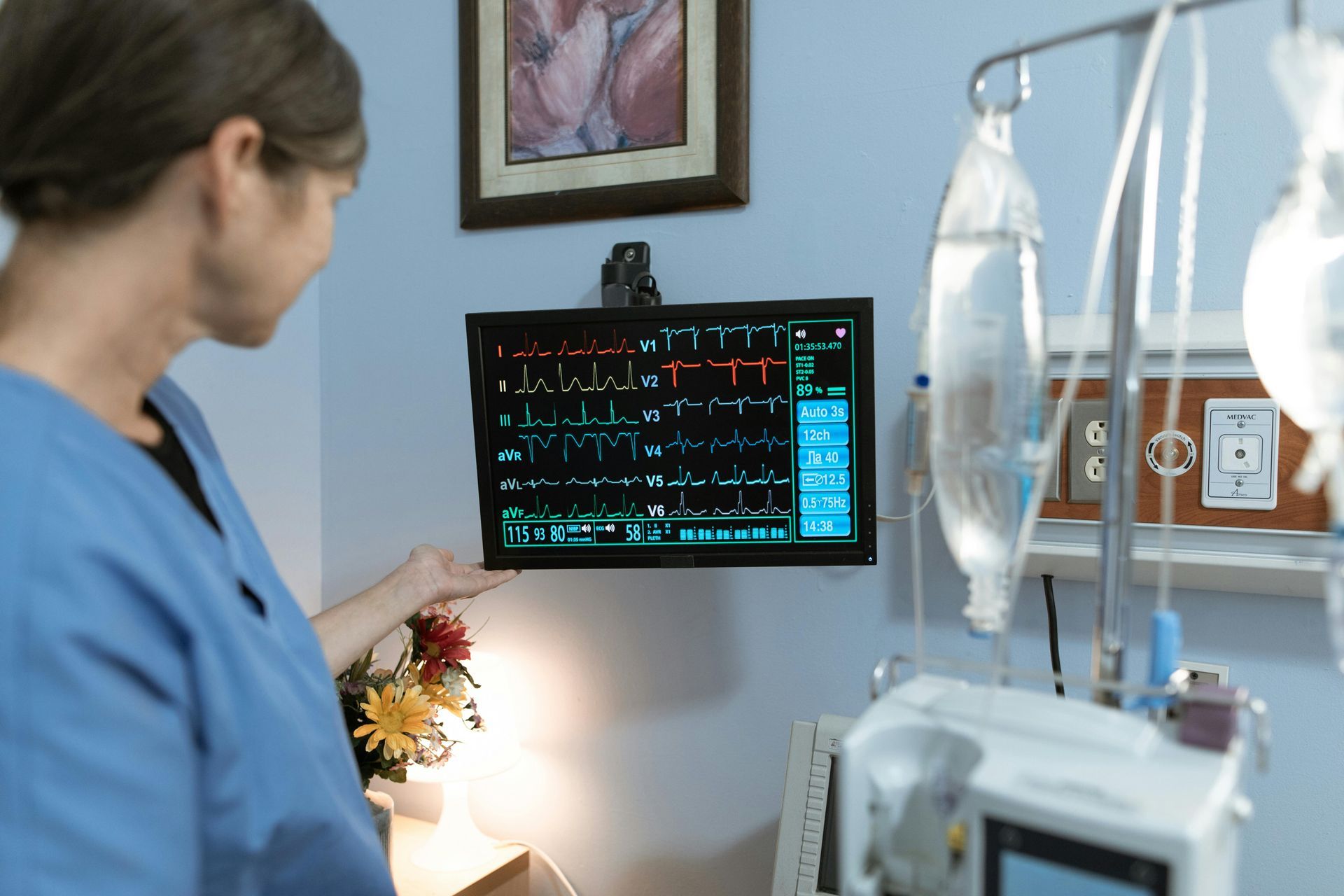Médico observando o monitor do paciente ao lado da cama em um quarto de hospital, próximo a bolsas de soro e equipamentos médicos.