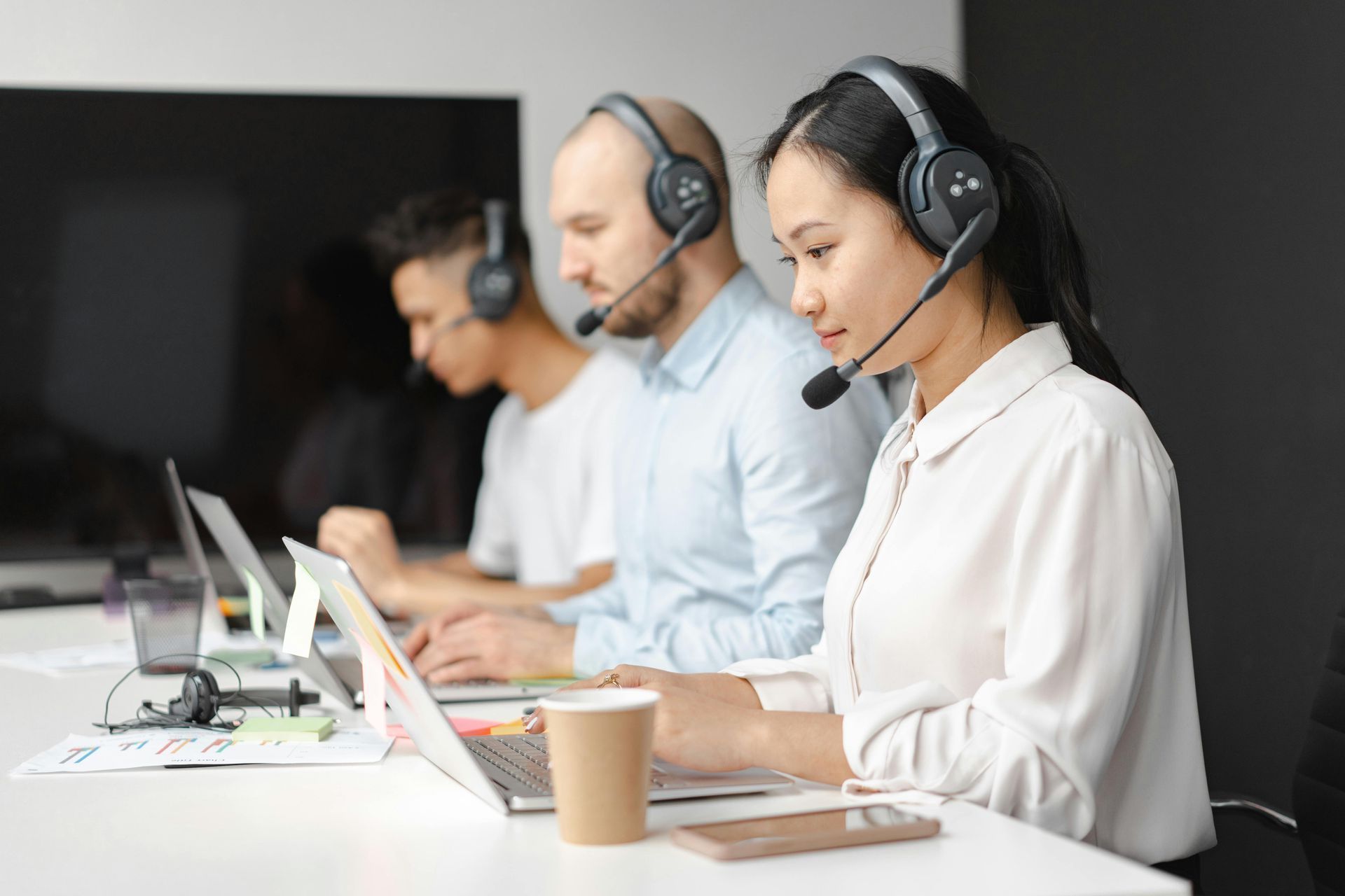 Equipe de suporte ao cliente usando fones de ouvido, trabalhando em laptops em um escritório bem iluminado.