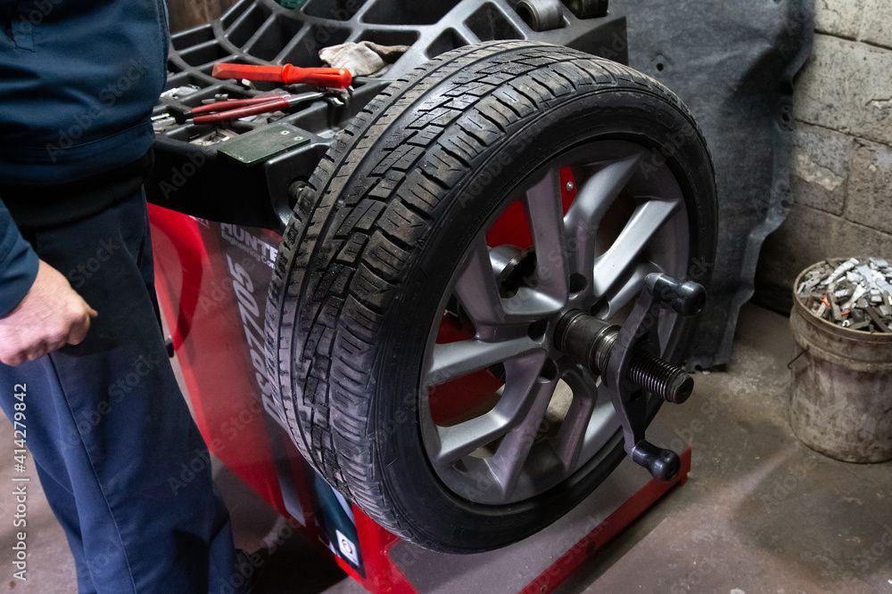 Tire being balanced on a red machine in a garage setting; a person's arm is visible on the left.