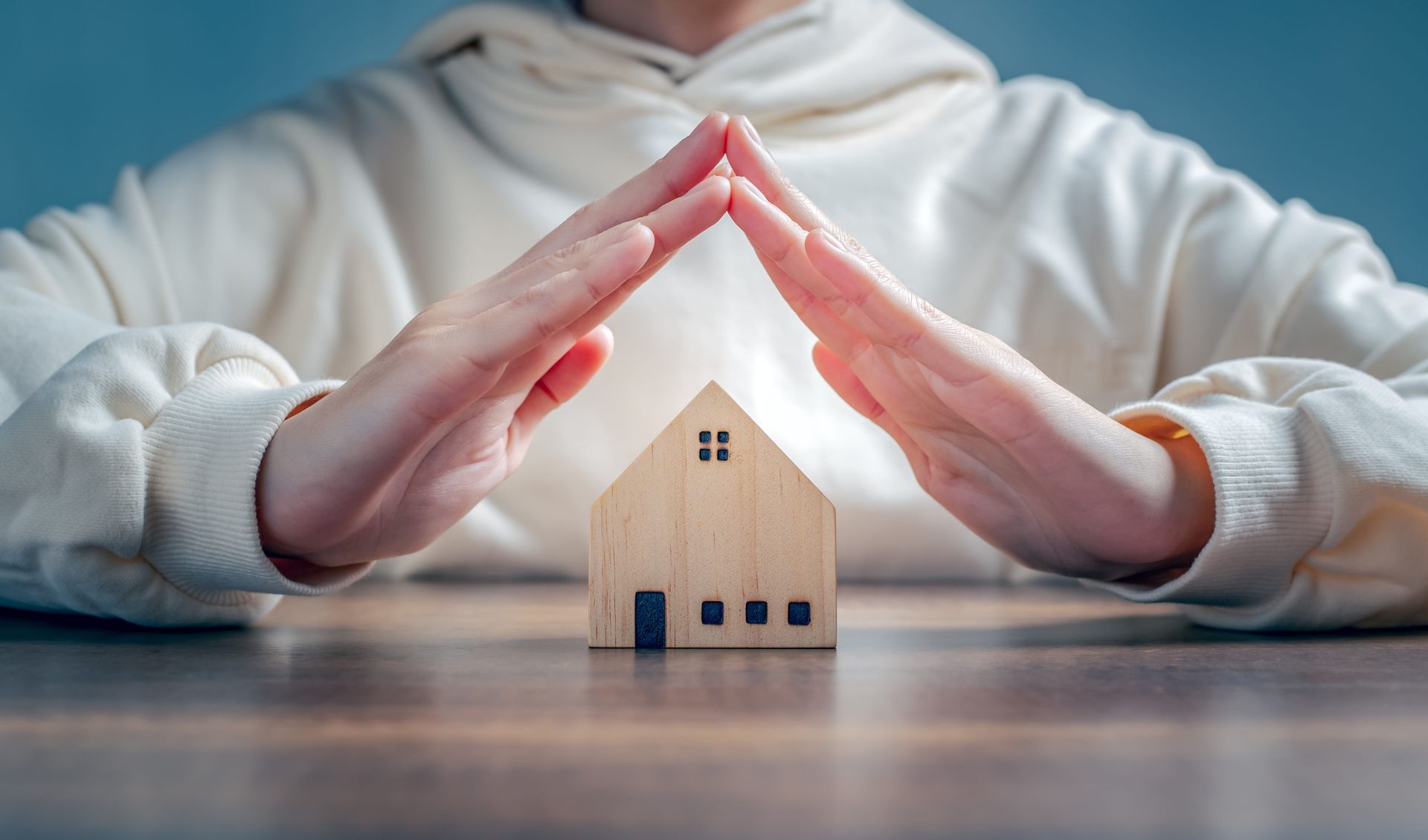 Hands forming a roof over a small wooden house model on a table