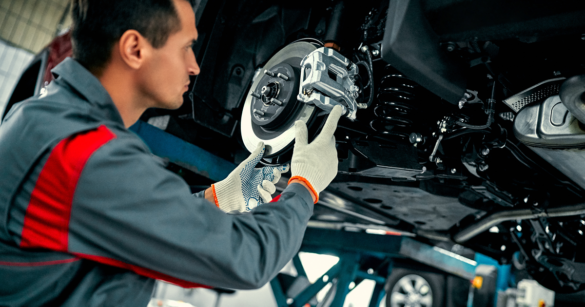 Mechanic working on car brakes; wearing gloves and gray uniform, inside a garage.
