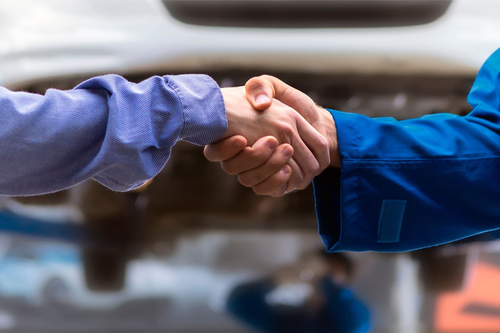 Two people shaking hands near a car; one in a blue mechanic's jumpsuit, the other in a blue shirt.