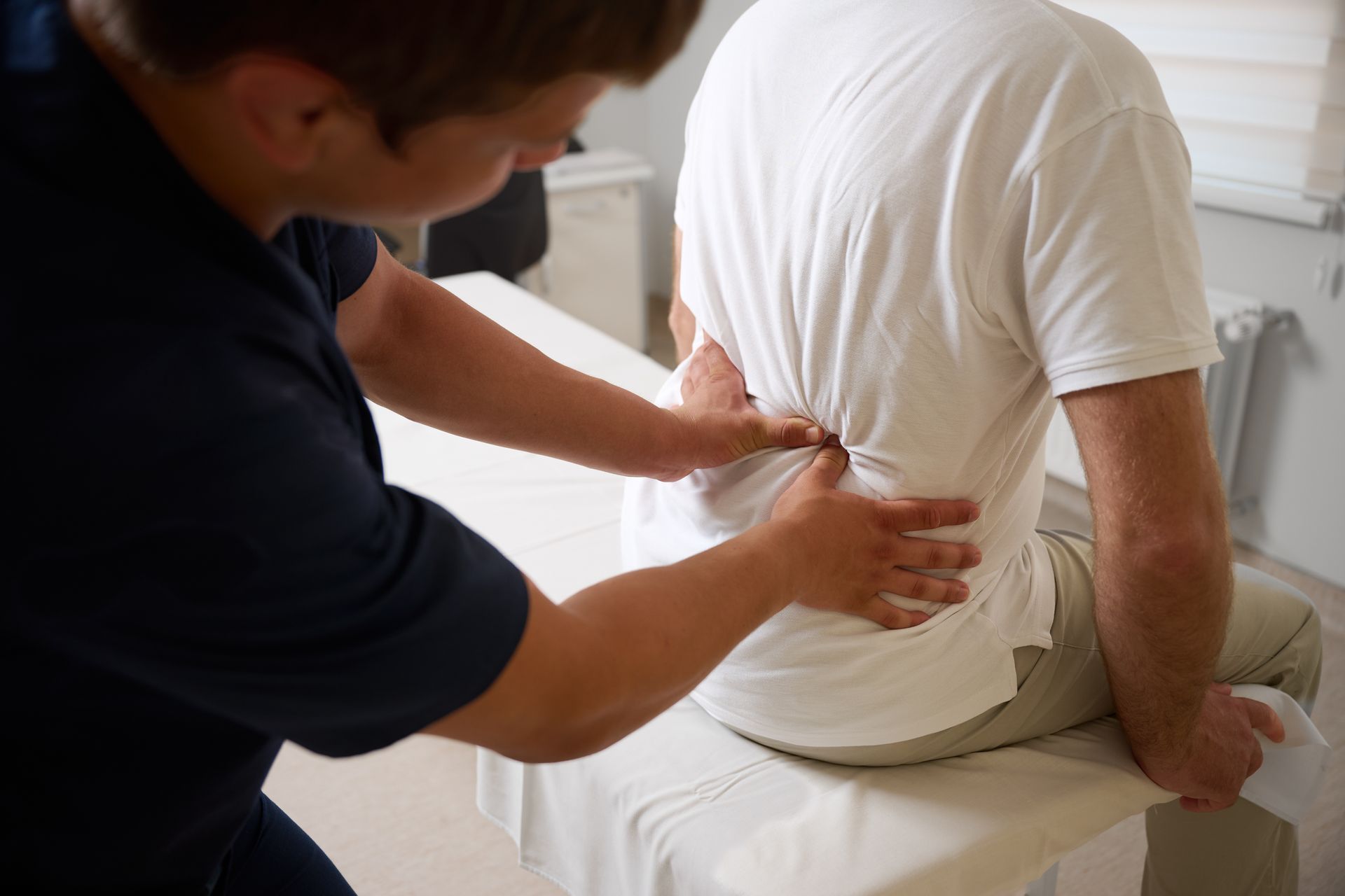 Person massaging another person's lower back while they sit on a bed in a bright room