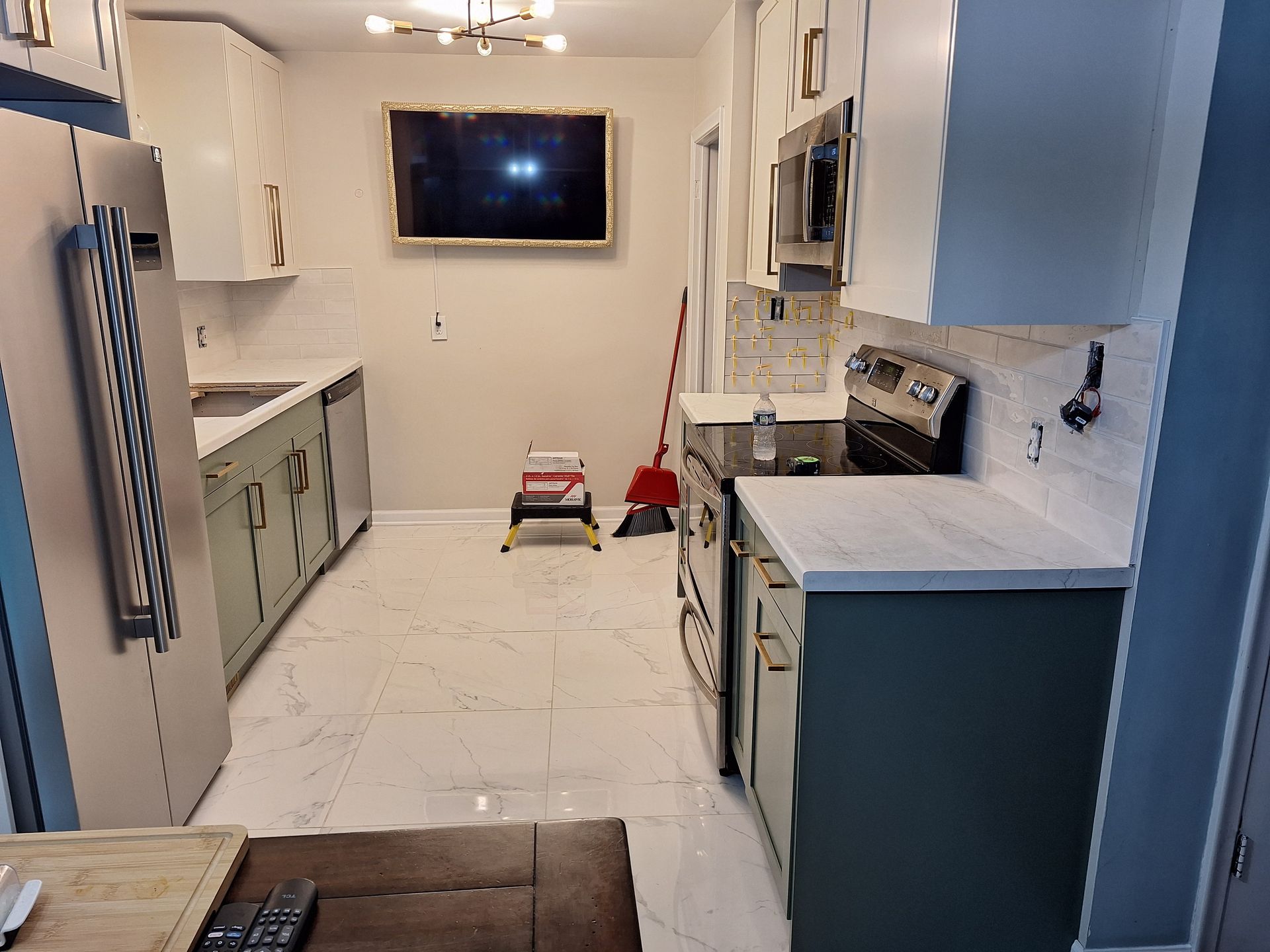Narrow white kitchen with blue cabinets, marble counters, wall TV, and a small step stool centered on the floor.
