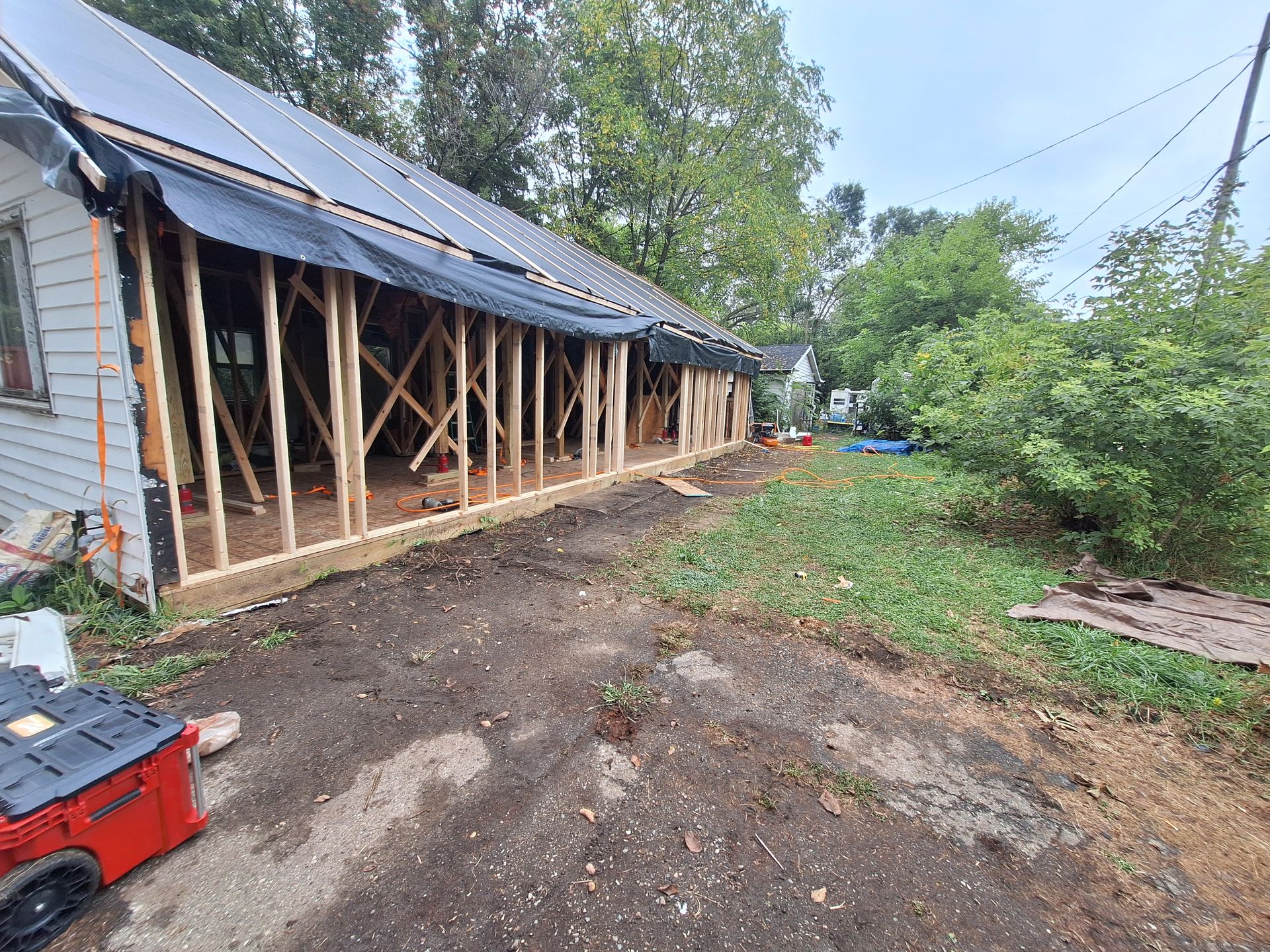 Partially rebuilt house with exposed framing, black tarp roof, and muddy yard beside trees