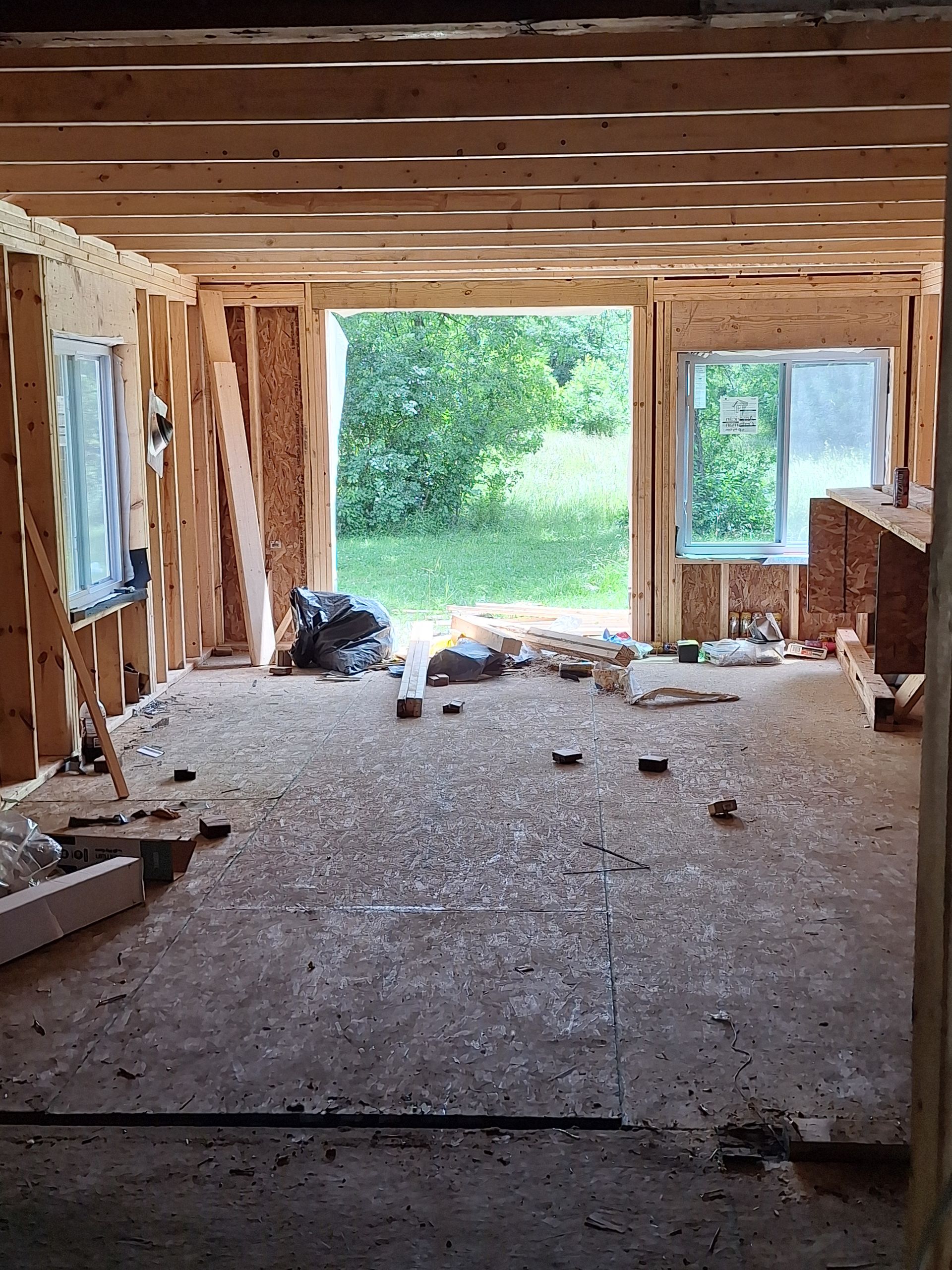 Unfinished room with exposed studs, debris on the floor, and open windows and doorway to greenery outside