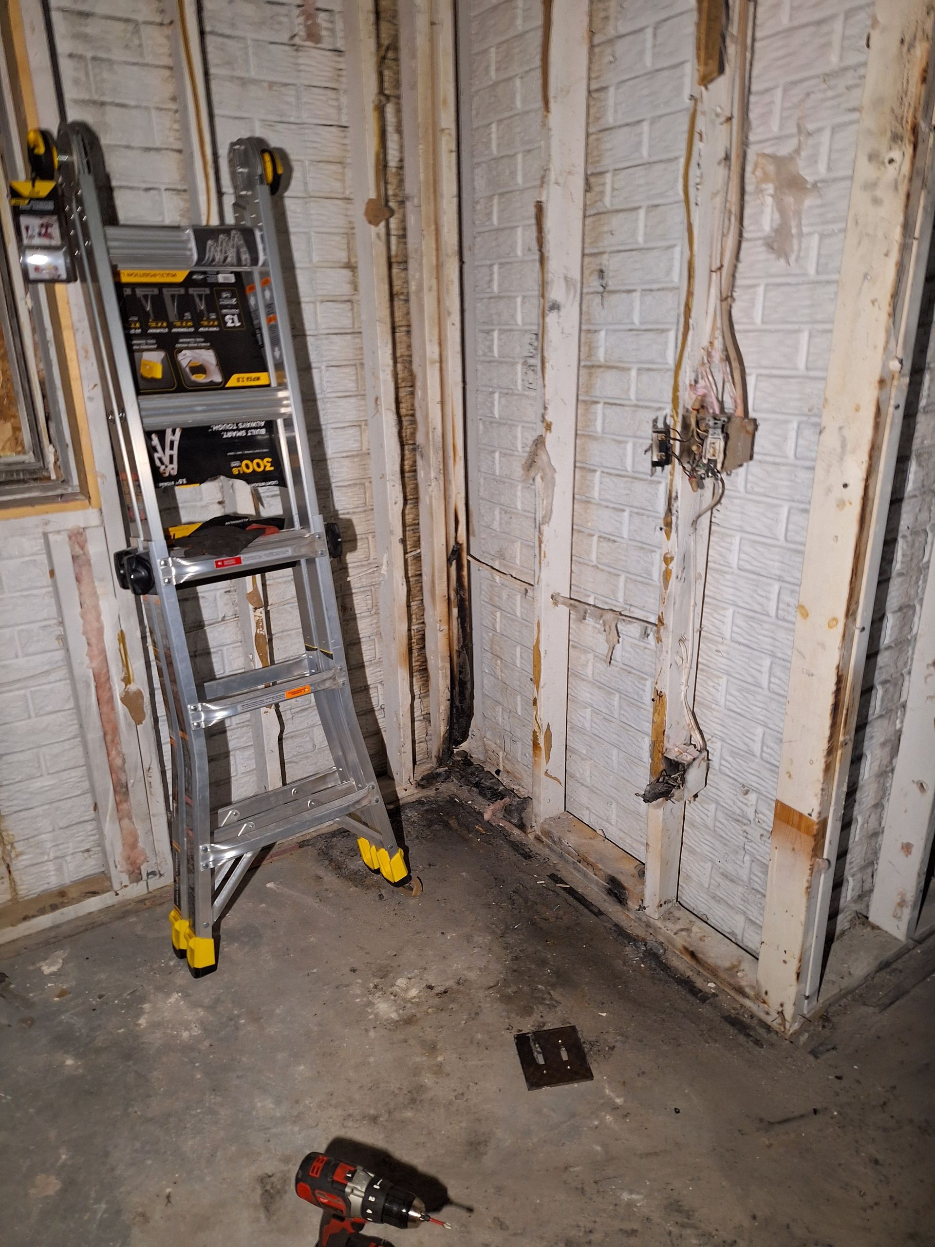 Ladder and tools in a bare, unfinished basement corner with white brick walls and concrete floor