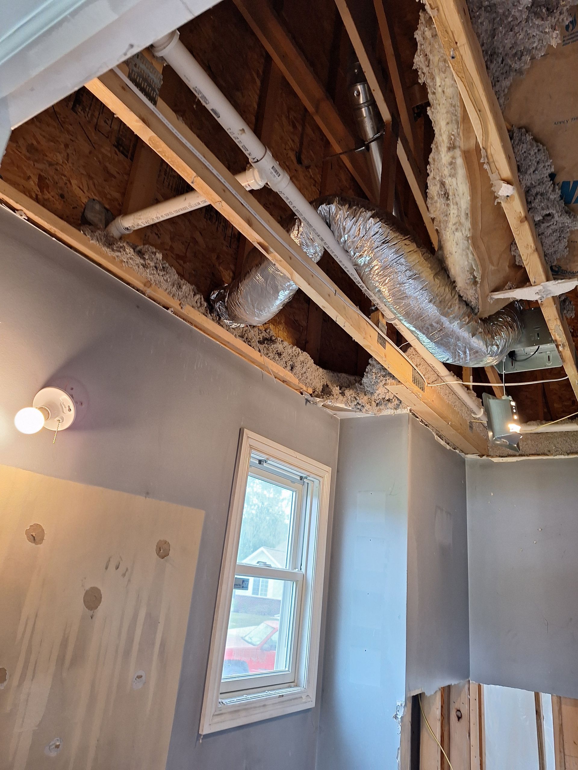 Partially demolished bathroom ceiling with exposed wooden joists, ductwork, and insulation above a window and wall.