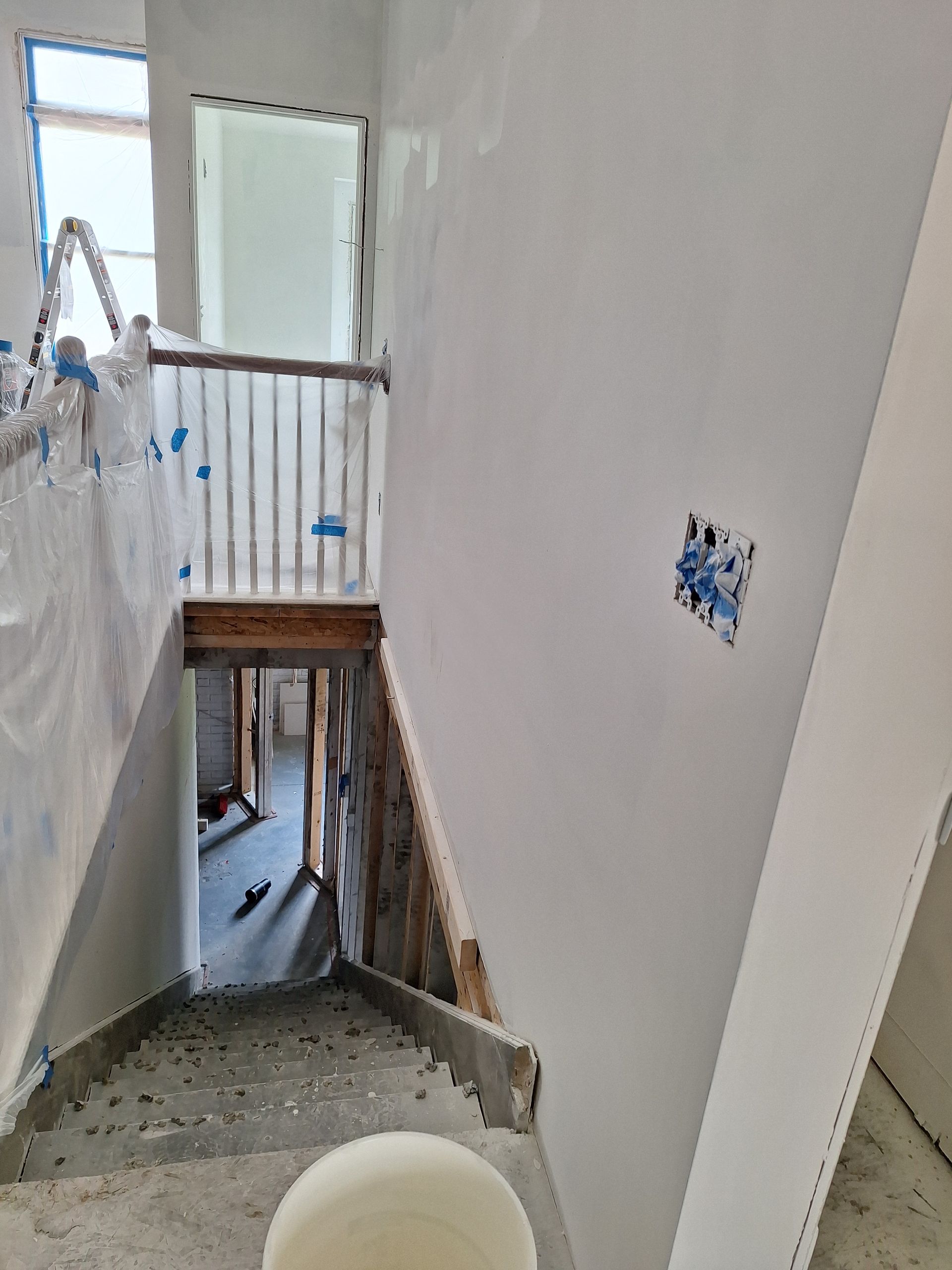 Stairwell under renovation with plastic sheeting, drywall, and a narrow opening to a lower level
