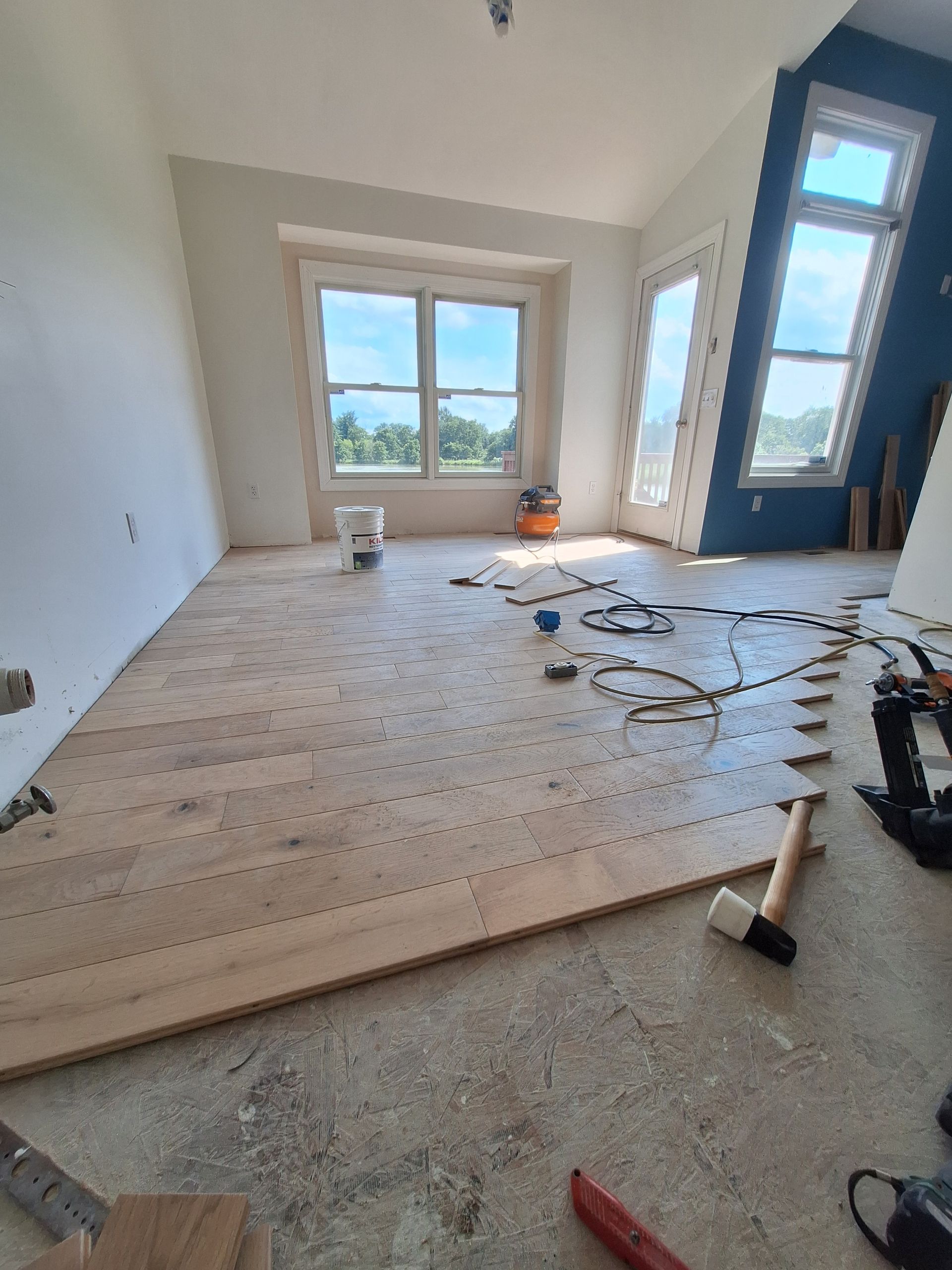 Empty room under renovation with exposed floorboards, tools, and large windows letting in daylight.
