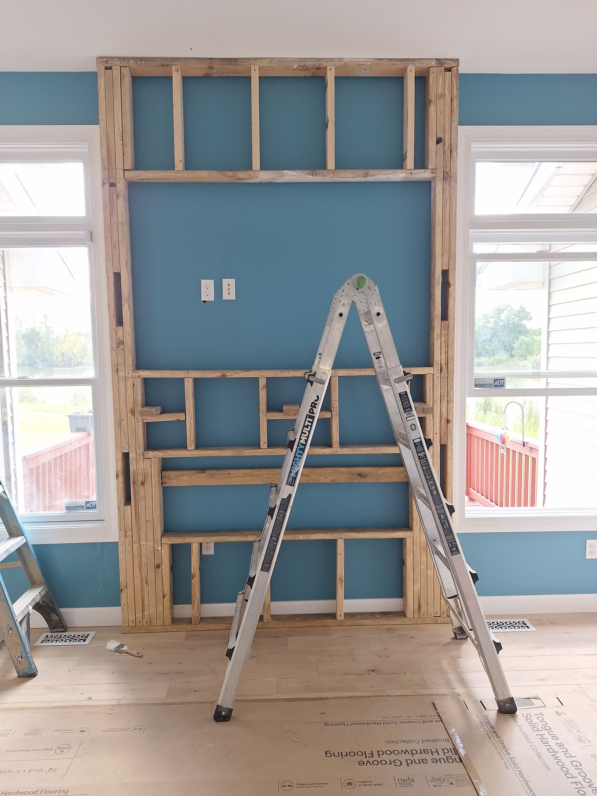 Ladders and wall framing in a room under renovation with blue walls and windows on both sides