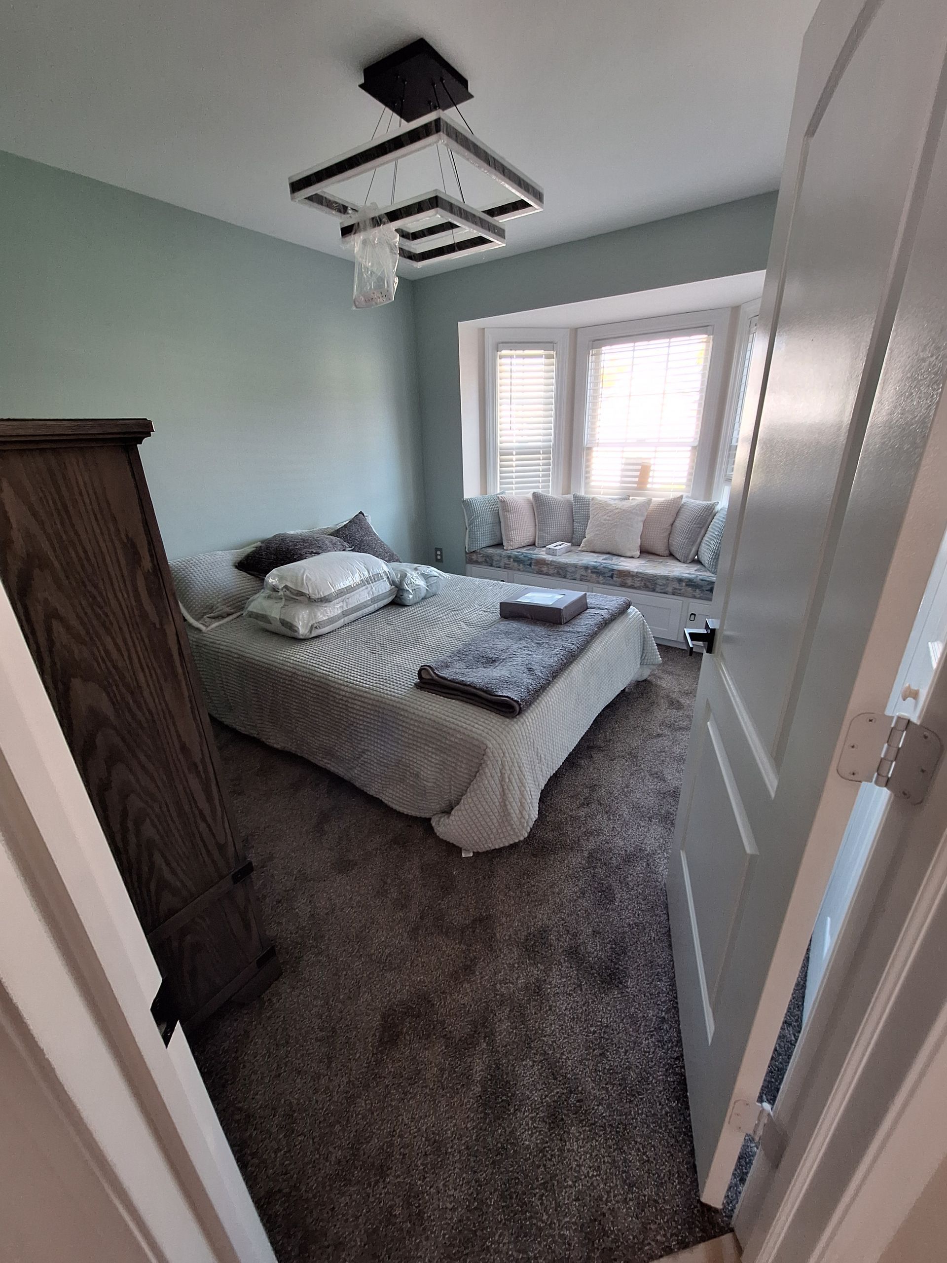 Bright bedroom with an unmade bed, gray carpet, white walls, and a window with blinds.