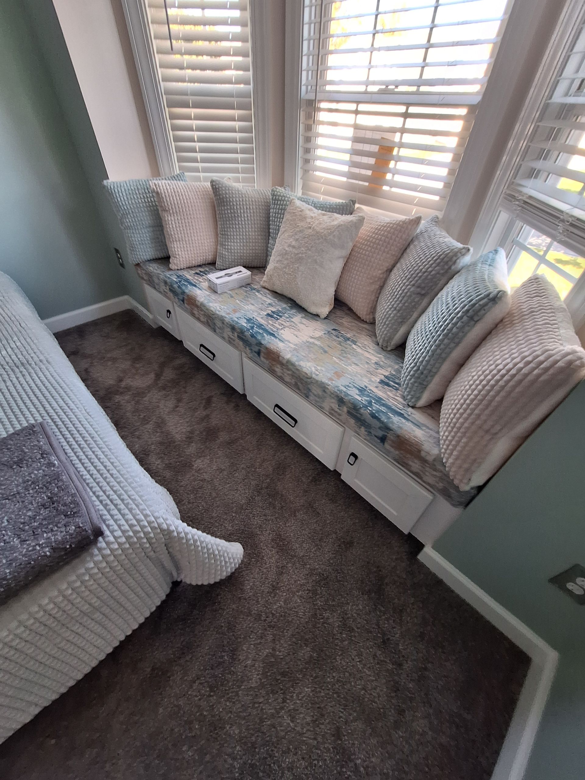 Bright bay window nook with a white cushioned bench, patterned pillows, and a gray carpet in a soft teal room