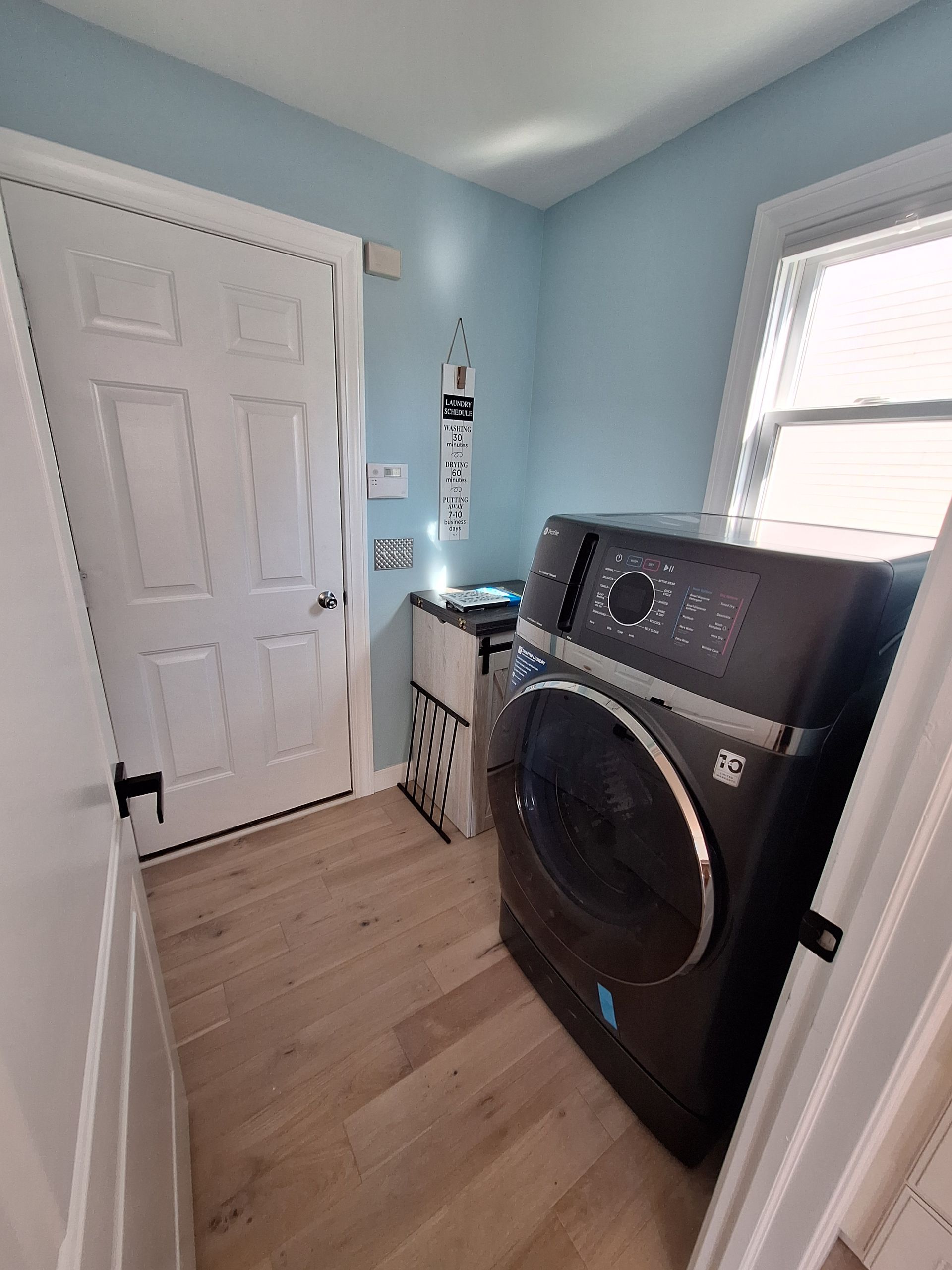 Small laundry room with light blue walls, white door, window, and a front-loading washer and dryer.