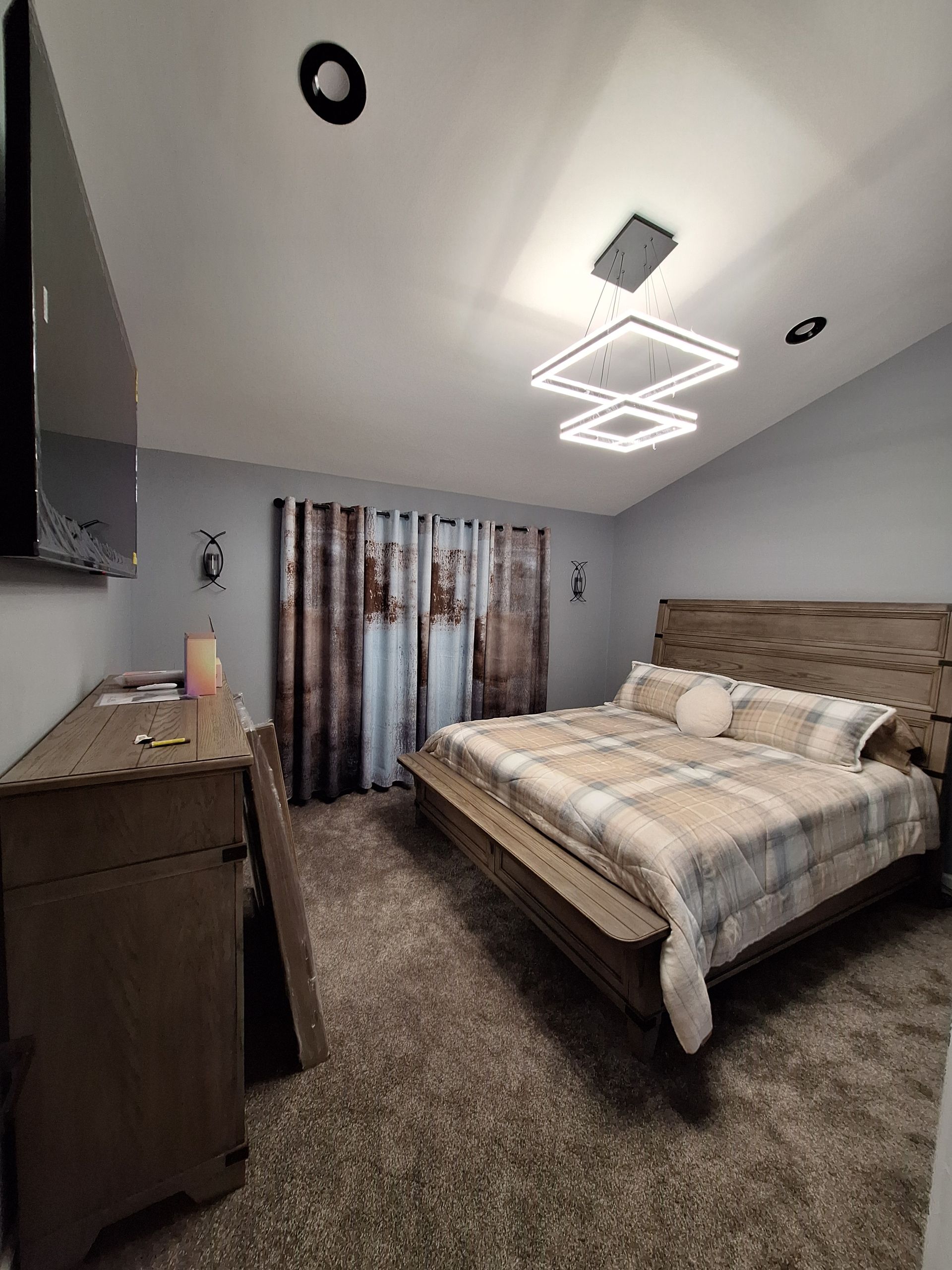 Bedroom with a large bed, dresser, and geometric ceiling light in a carpeted room.