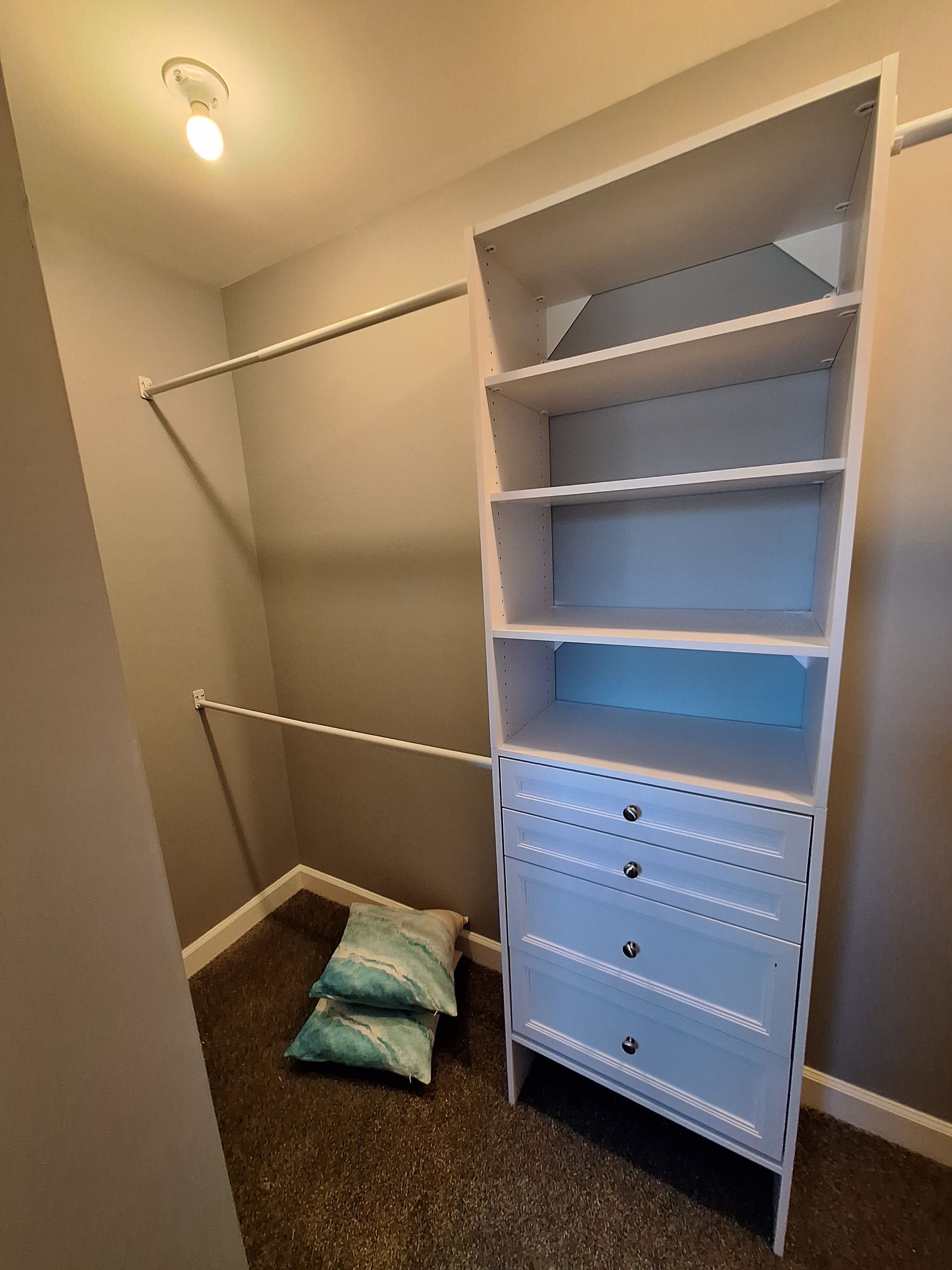 Small walk-in closet with shelves, hanging rods, and a white drawer unit; pillows sit on the floor.