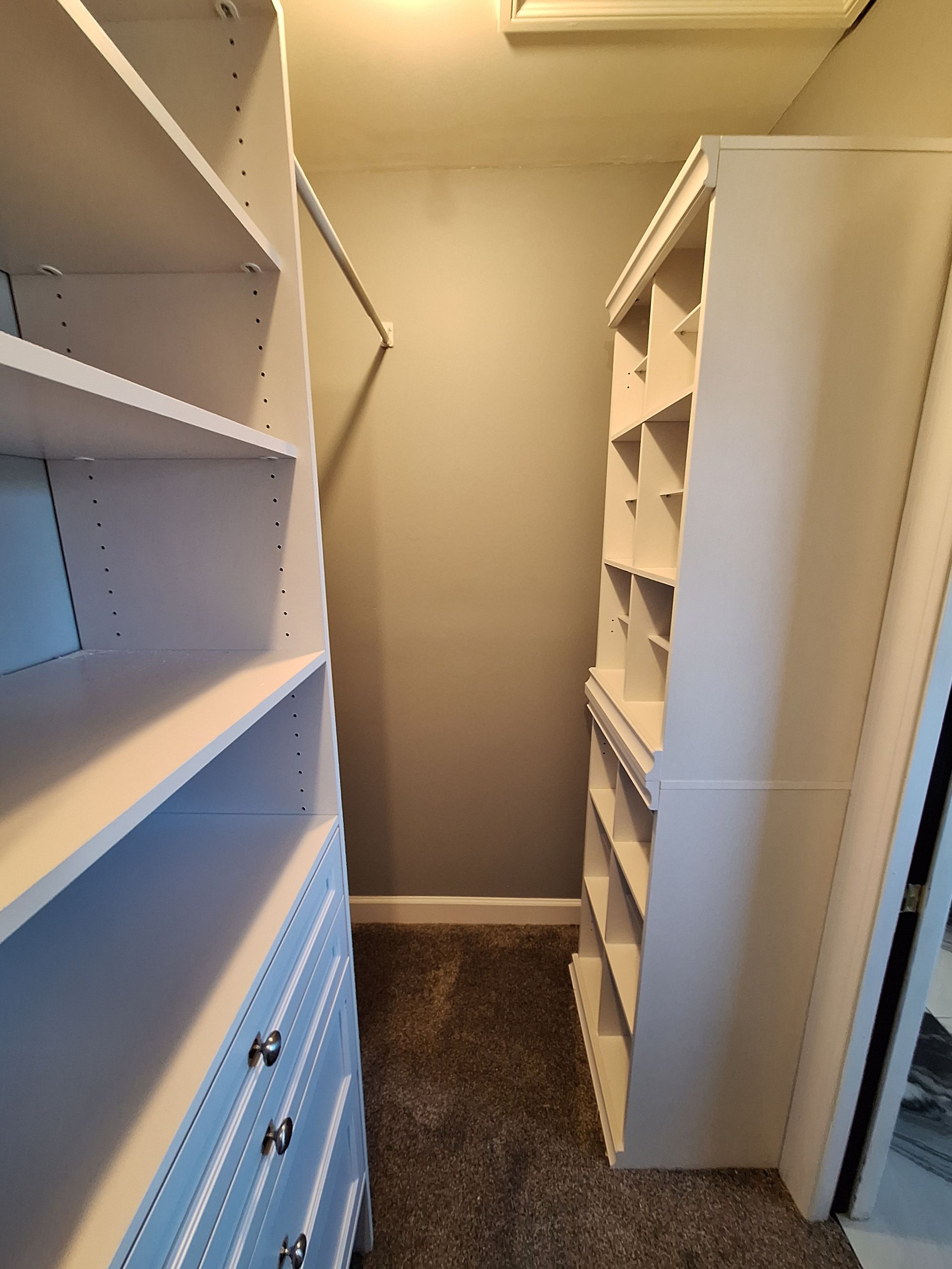 Narrow walk-in closet with white shelving and drawers on both sides and carpeted floor.