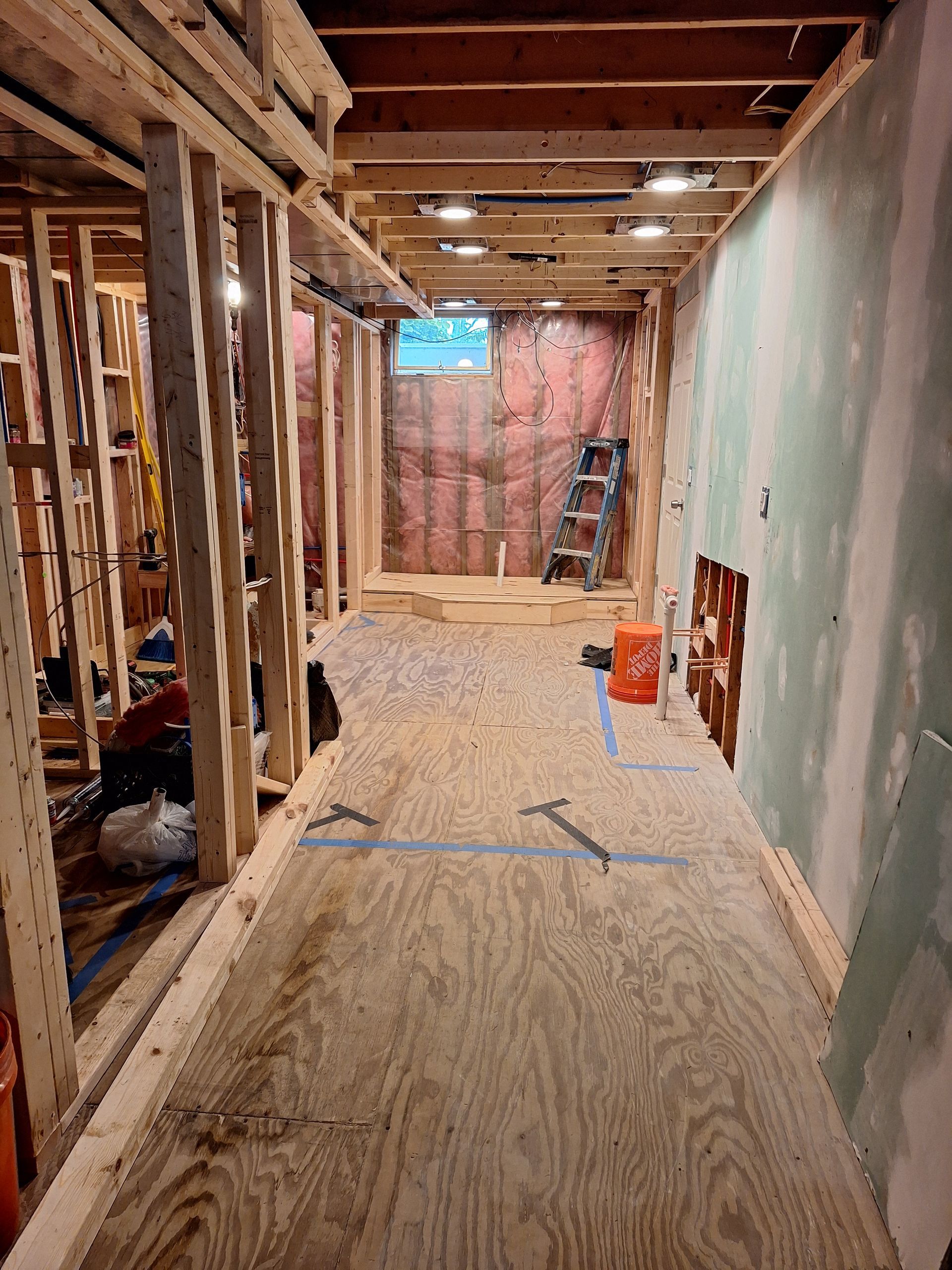 Interior under renovation with exposed studs, plywood floor, and ladder near the back wall
