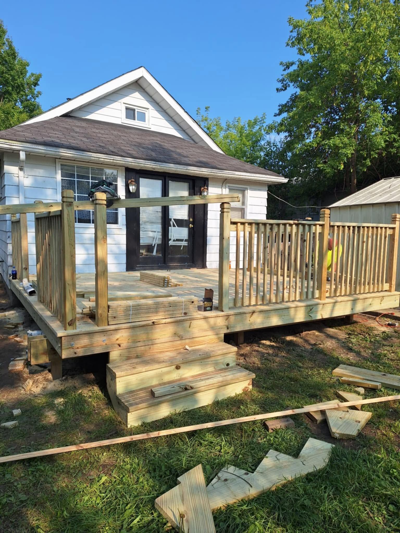 Weathered wooden deck with steps beside a house, surrounded by trees and yard debris.