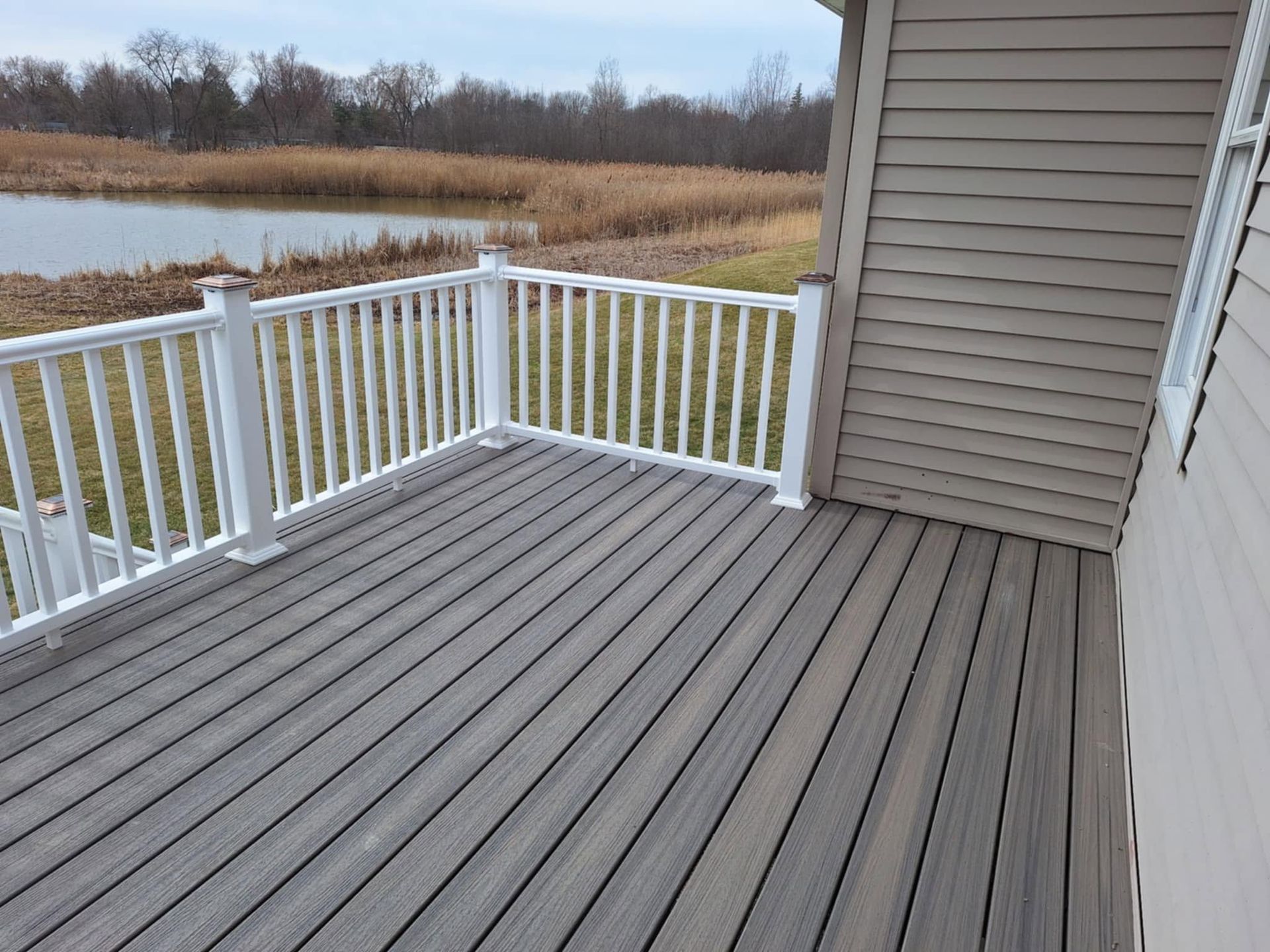 Empty gray deck with white railing beside a house, overlooking a marshy pond.