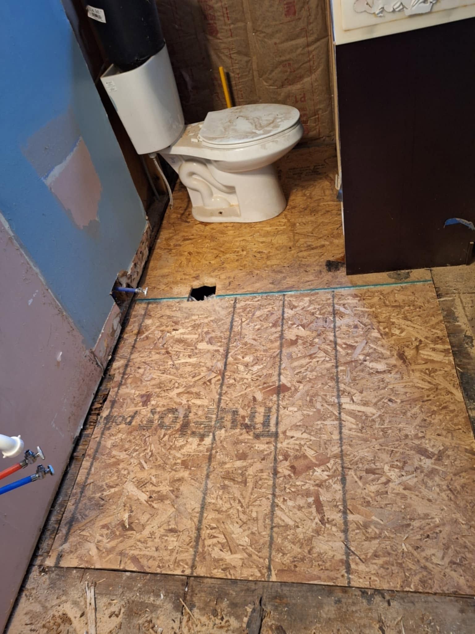 Small bathroom with a toilet, damaged plywood floor, and a visible hole near the doorway.