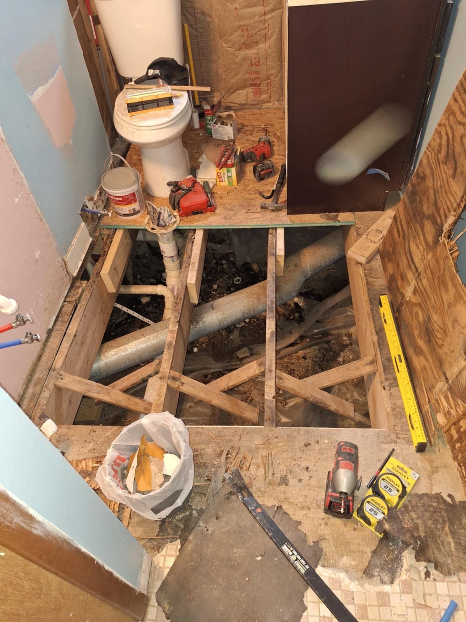 Bathroom floor under renovation, with exposed joists, tools, debris, and a toilet nearby.