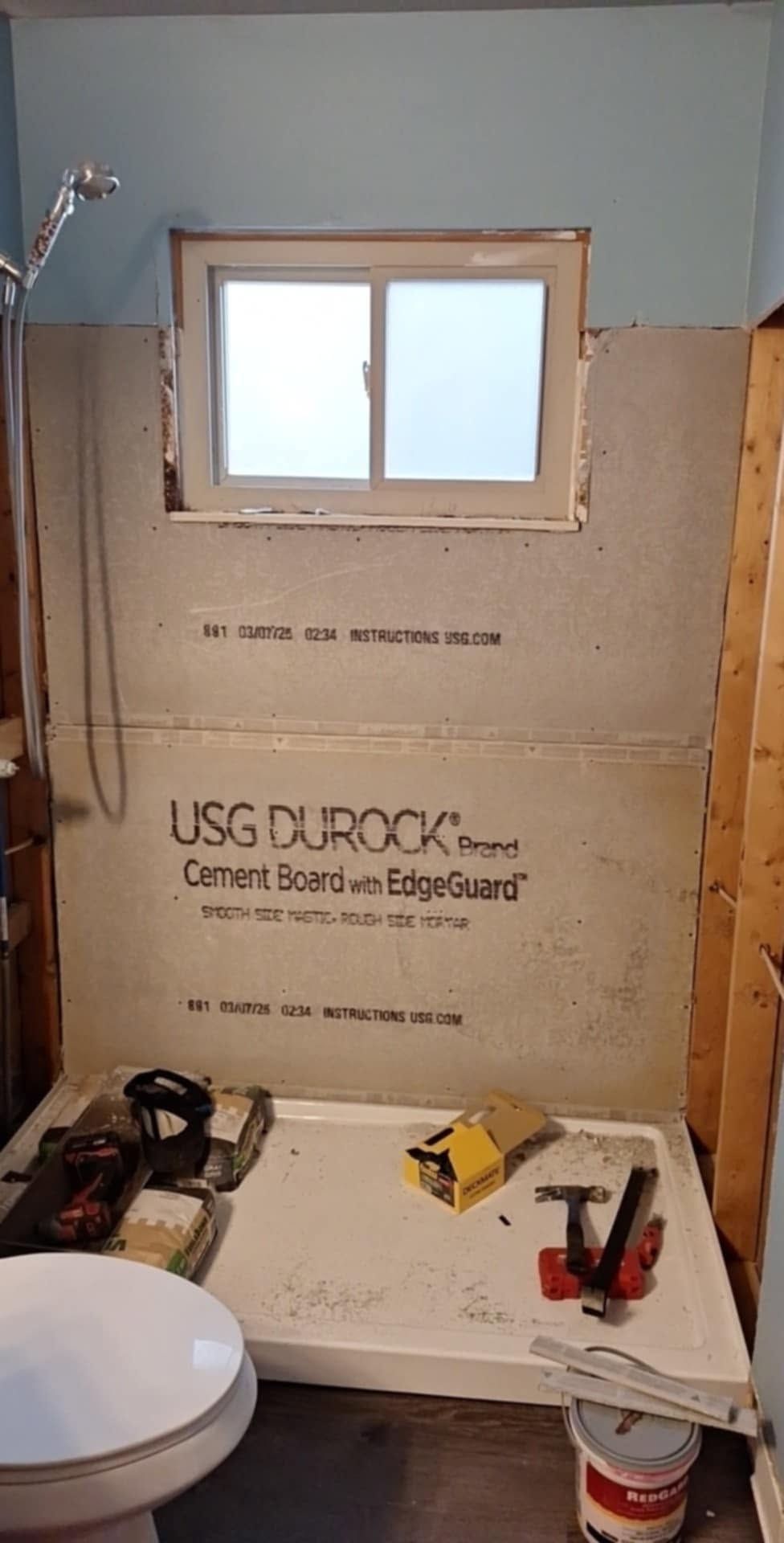 Bathroom under renovation with exposed shower wall, window, tools, and toilet visible