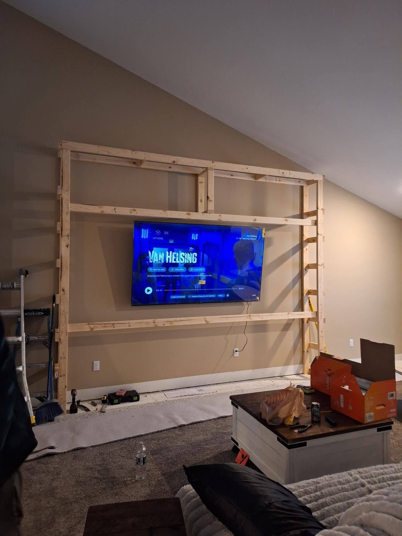 TV mounted in a wooden frame on an unfinished living room wall with tools and boxes nearby