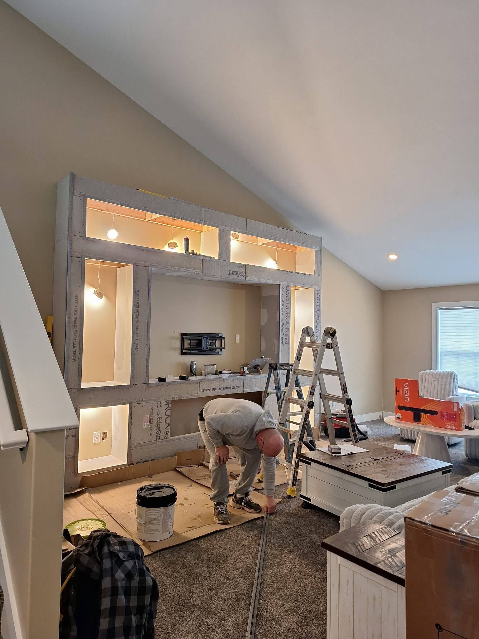 Person assembling a built-in wall unit in a living room with ladders, tools, and exposed wiring