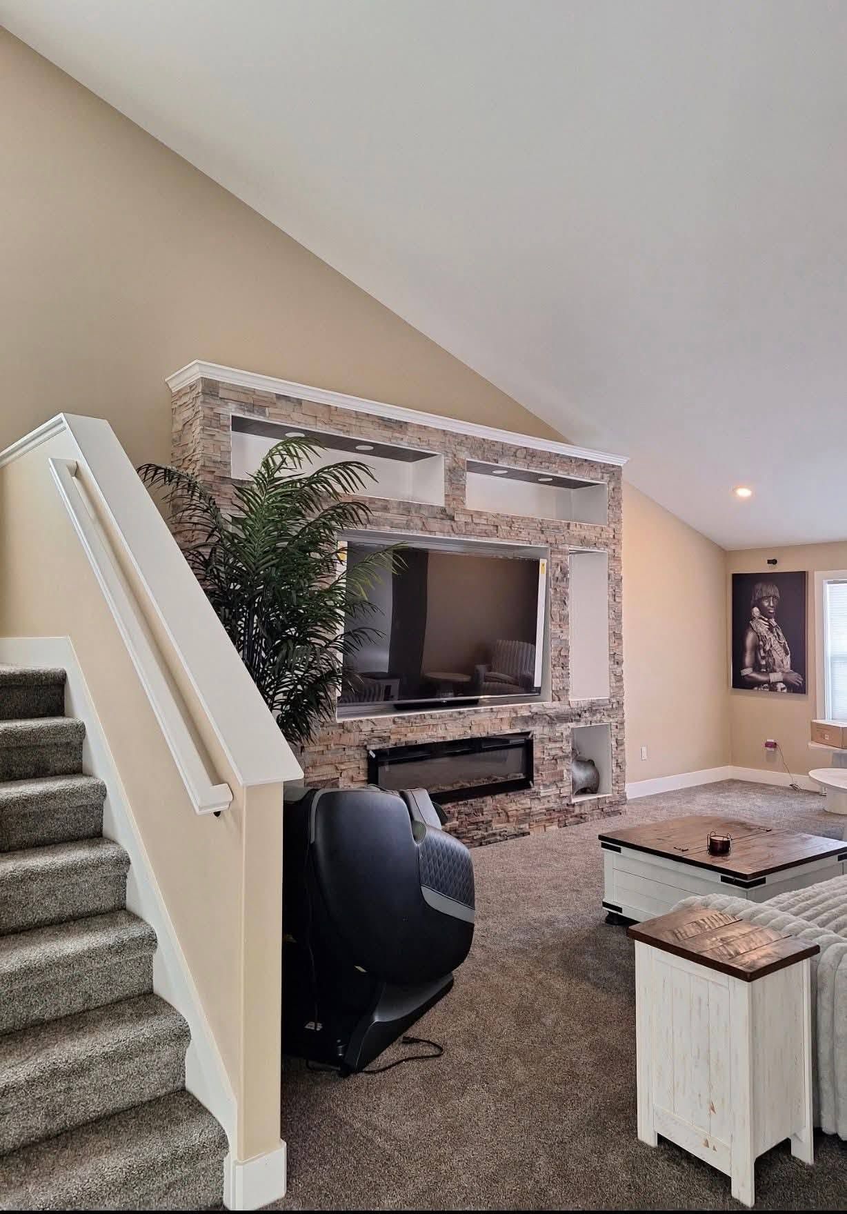 Carpeted living room with stone fireplace, TV, staircase, recliner, and large window.