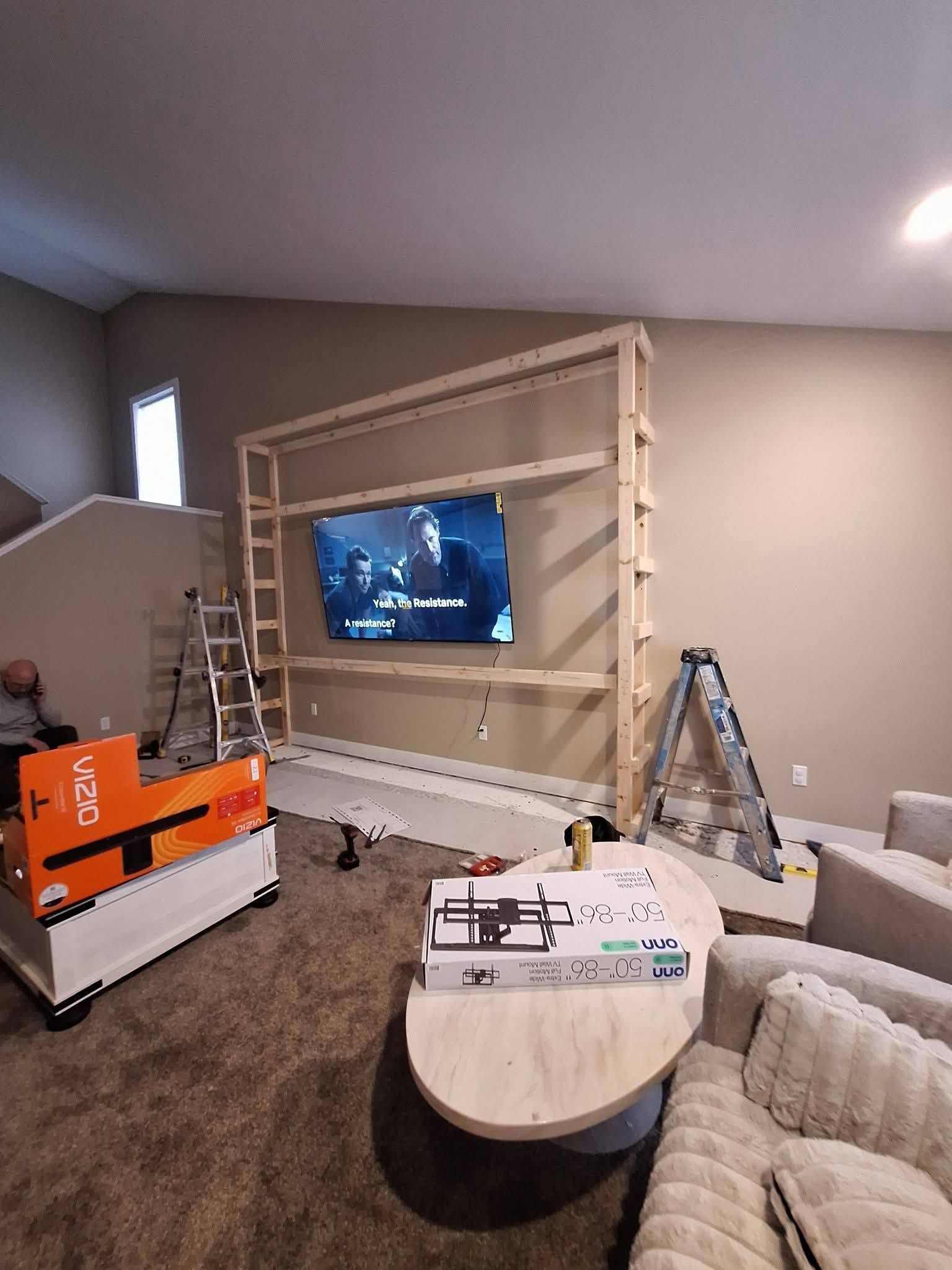 Living room under renovation with ladders, tools, and a TV mounted on a partially built wall.