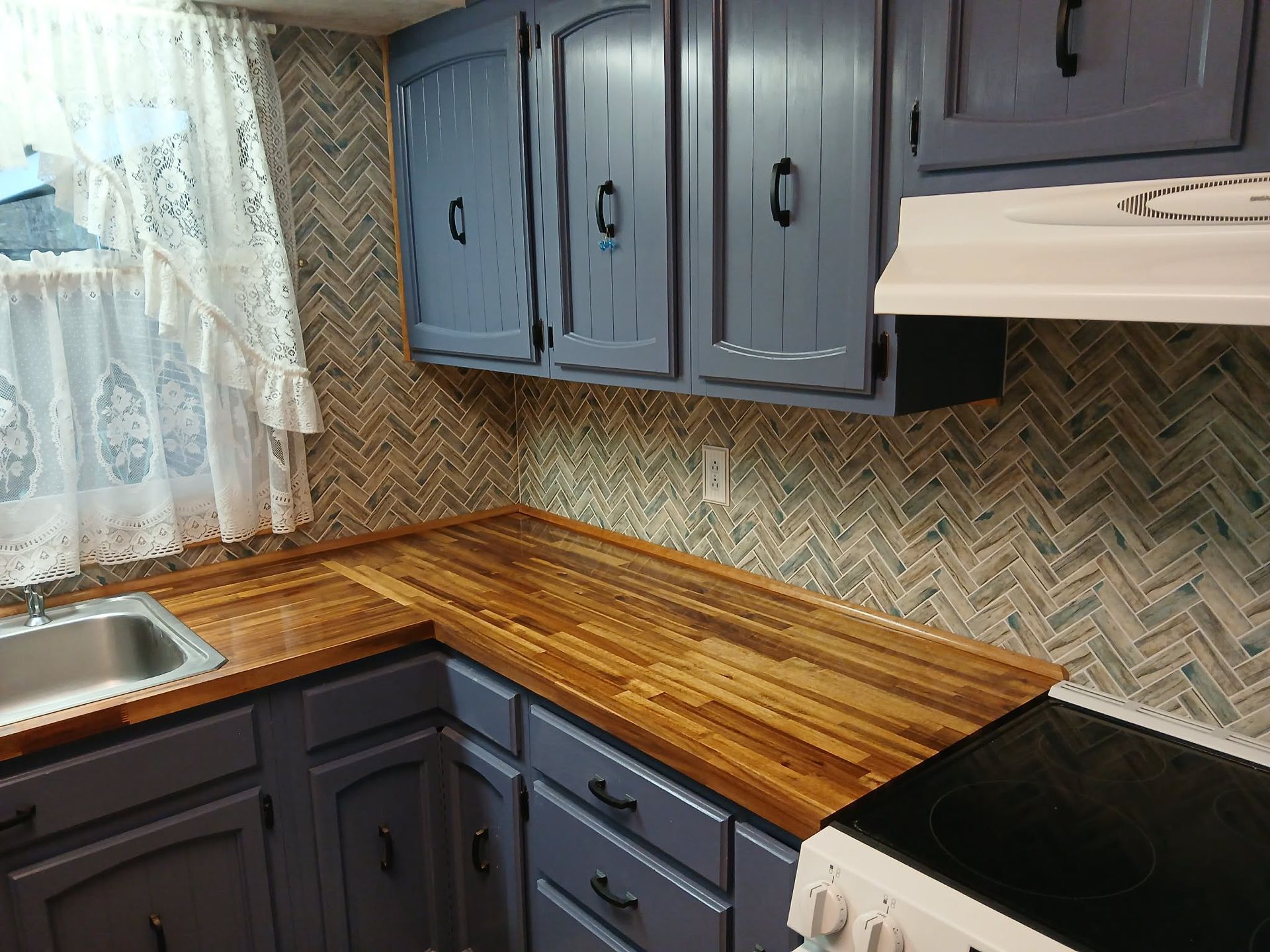 Small kitchen corner with dark cabinets, wood countertop, sink, and black stovetop under a herringbone backsplash.