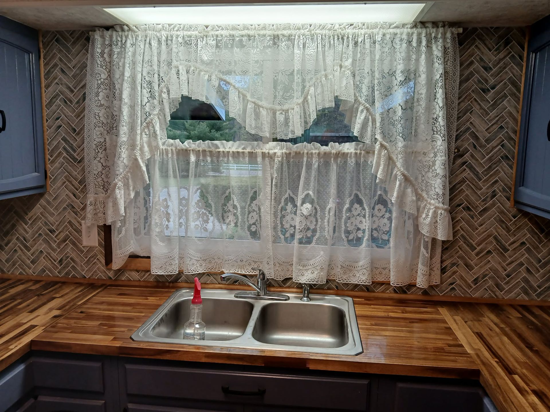 Kitchen sink beneath lace curtains and wooden countertops with blue cabinets.