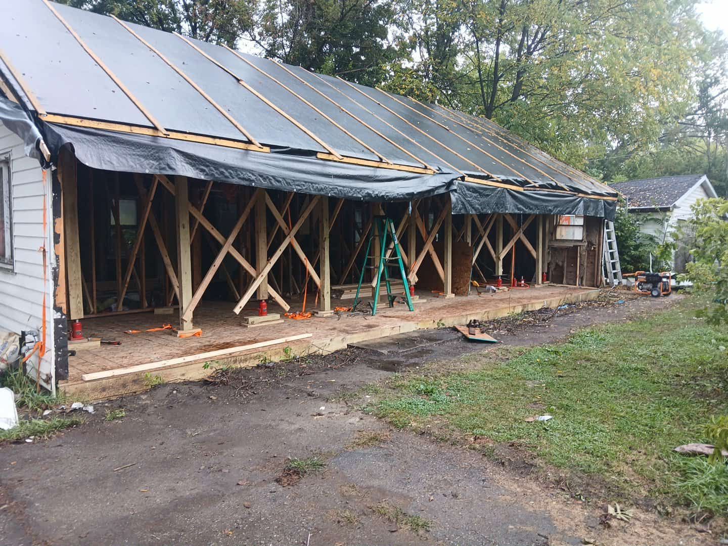 Partially demolished house with exposed framing, ladder, and solar panels on the roof, beside a dirt driveway