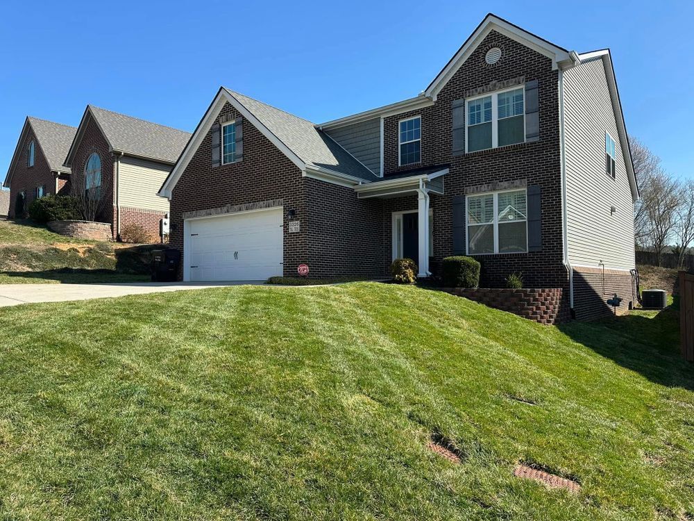 Two-story brick house on a slight hill with green grass. Another house is visible in the background.