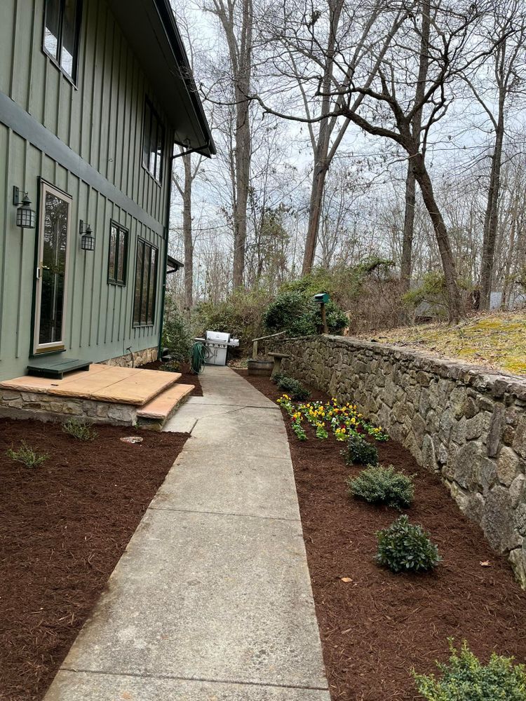 Side of a green house with a concrete walkway and stone wall, mulched flower beds, and trees.