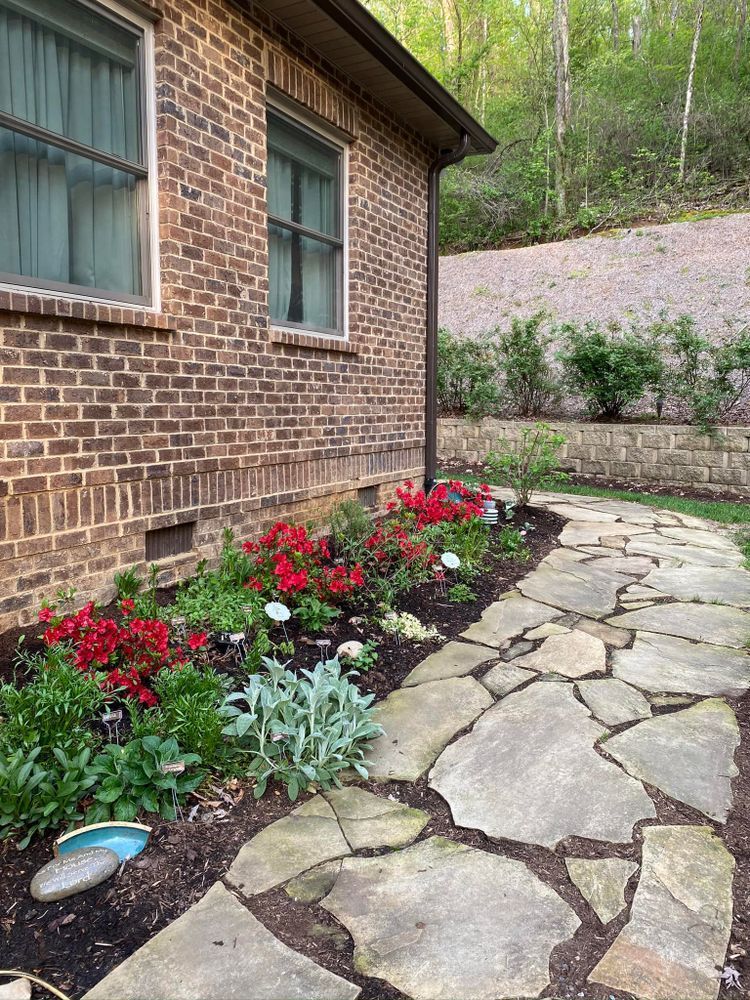Stone path curves past a brick house with a flower bed of red and green plants.