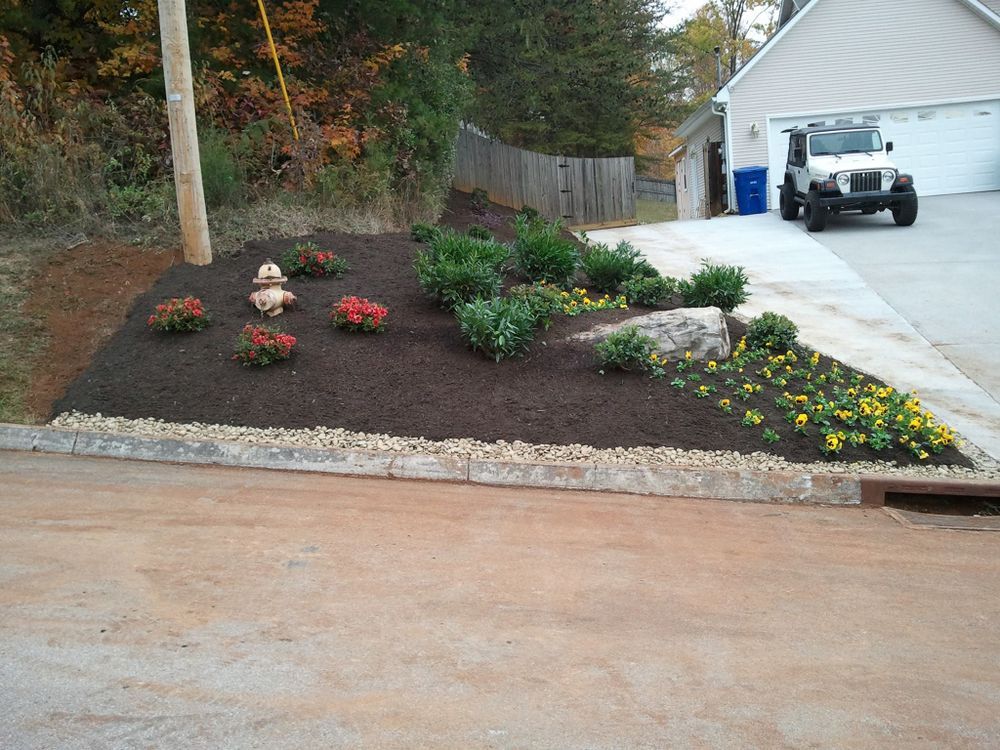 Flowerbed with various plants, bordered by stone and mulch, next to a driveway with a jeep.