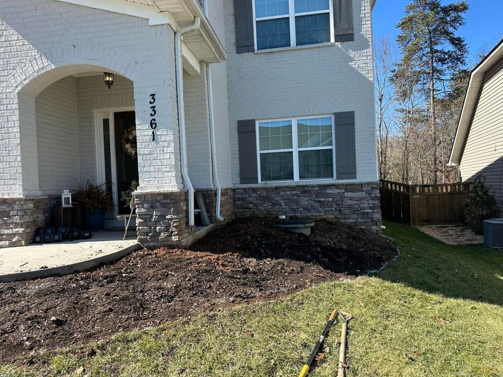 House exterior with brick, gray siding, and a mulched flower bed, tools in the grass.