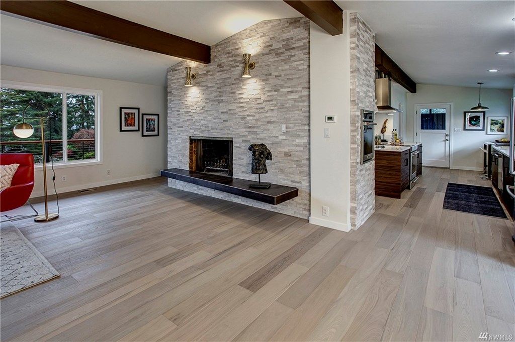 Spacious living room with stone fireplace, wood beams, pale floors, and large windows overlooking greenery