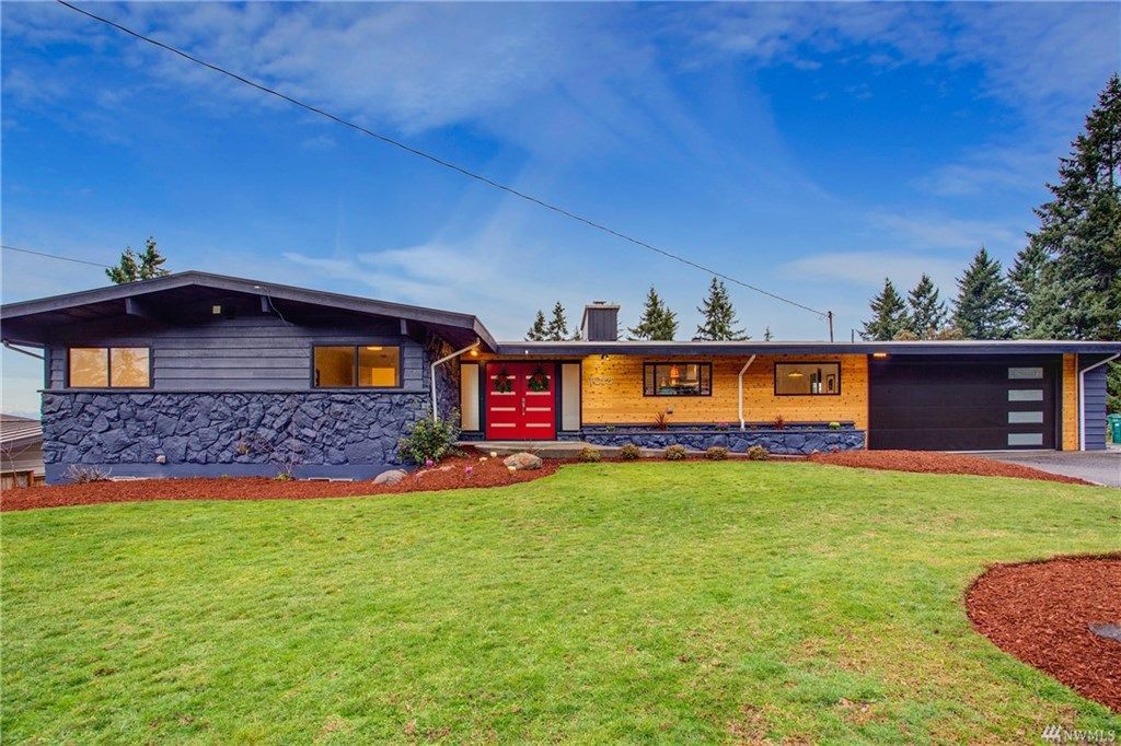 Modern single-story house with stone exterior, red front door, and large green lawn under a blue sky
