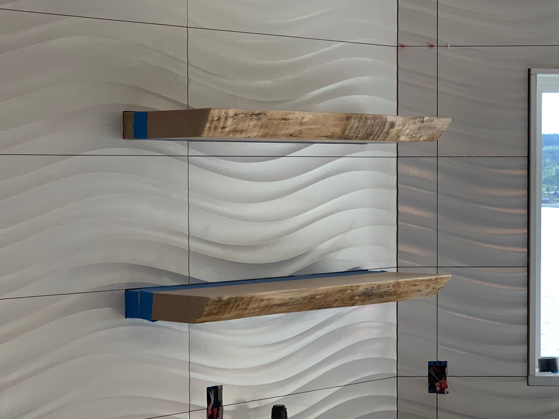 Two wall-mounted wooden shelves with blue supports on a white wavy wall near a window.