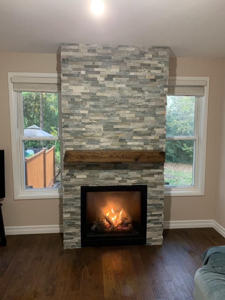 Living room fireplace with stacked stone surround, lit fire, two windows, and hardwood floor