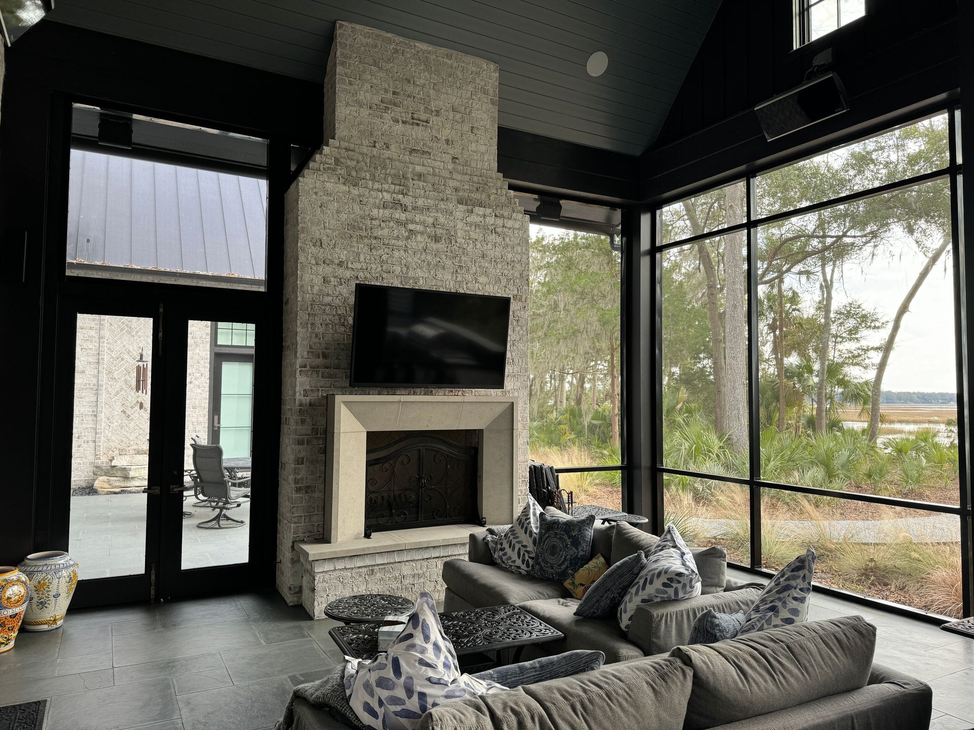 Modern living room with stone fireplace, dark walls, and large windows overlooking trees.